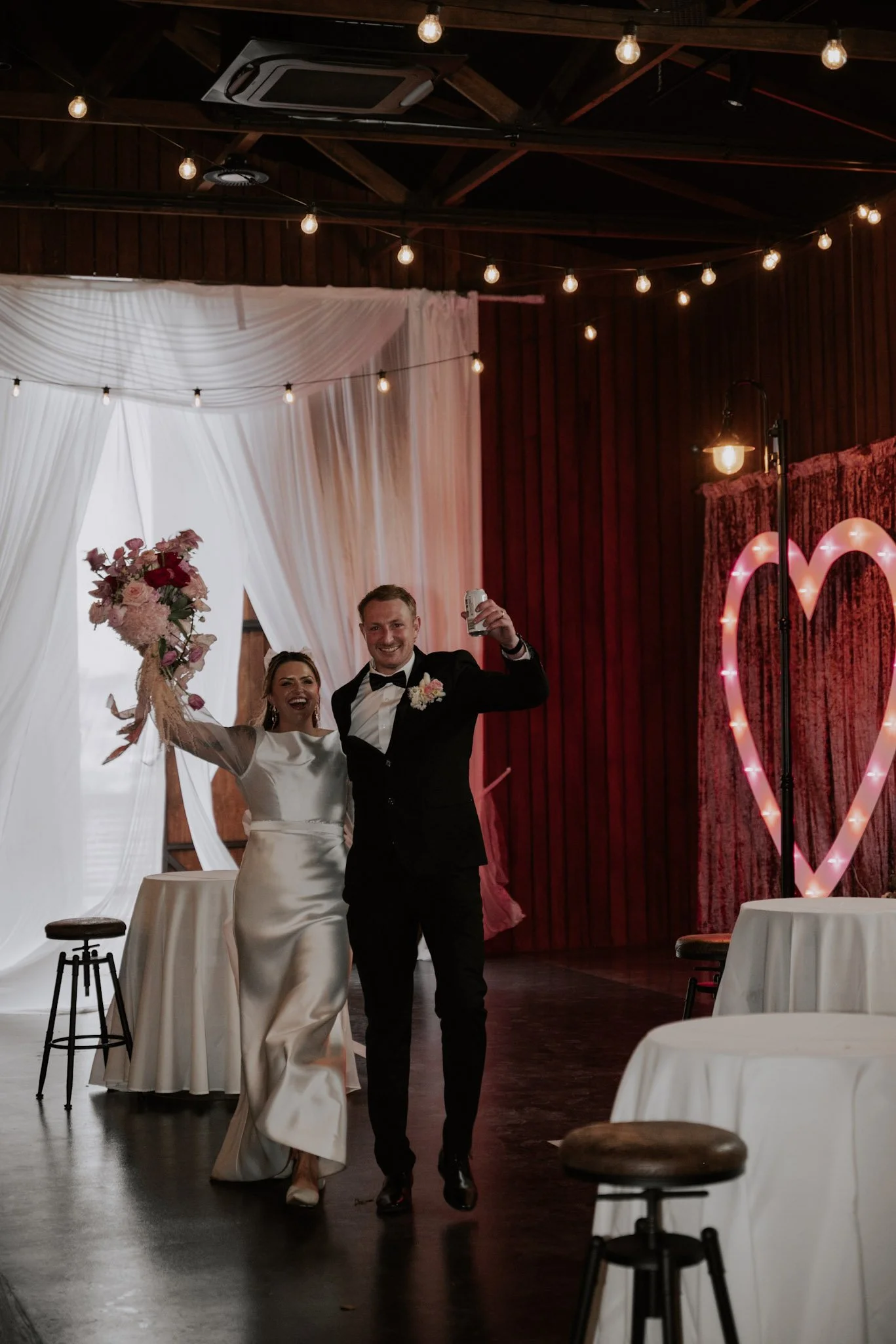 Bride and groom celebrating in a decorated wedding venue with string lights, a pink heart-shaped neon sign, and white drapes, with the bride holding a bouquet and both smiling.