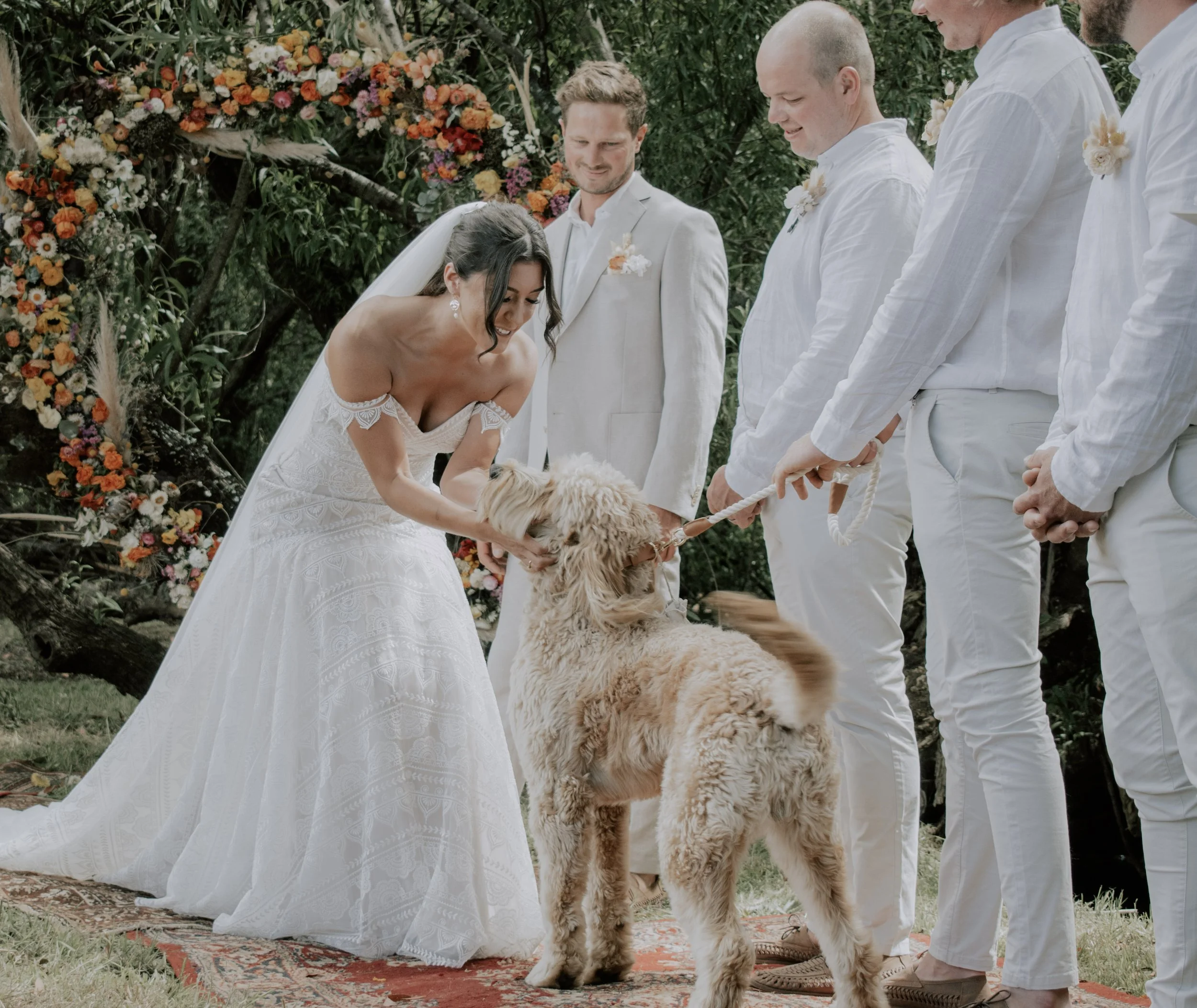Bride in a white wedding gown touching a golden retriever dog during outdoor wedding ceremony with bridesmaids and groomsmen, floral arch backdrop, on a decorative rug on grass.