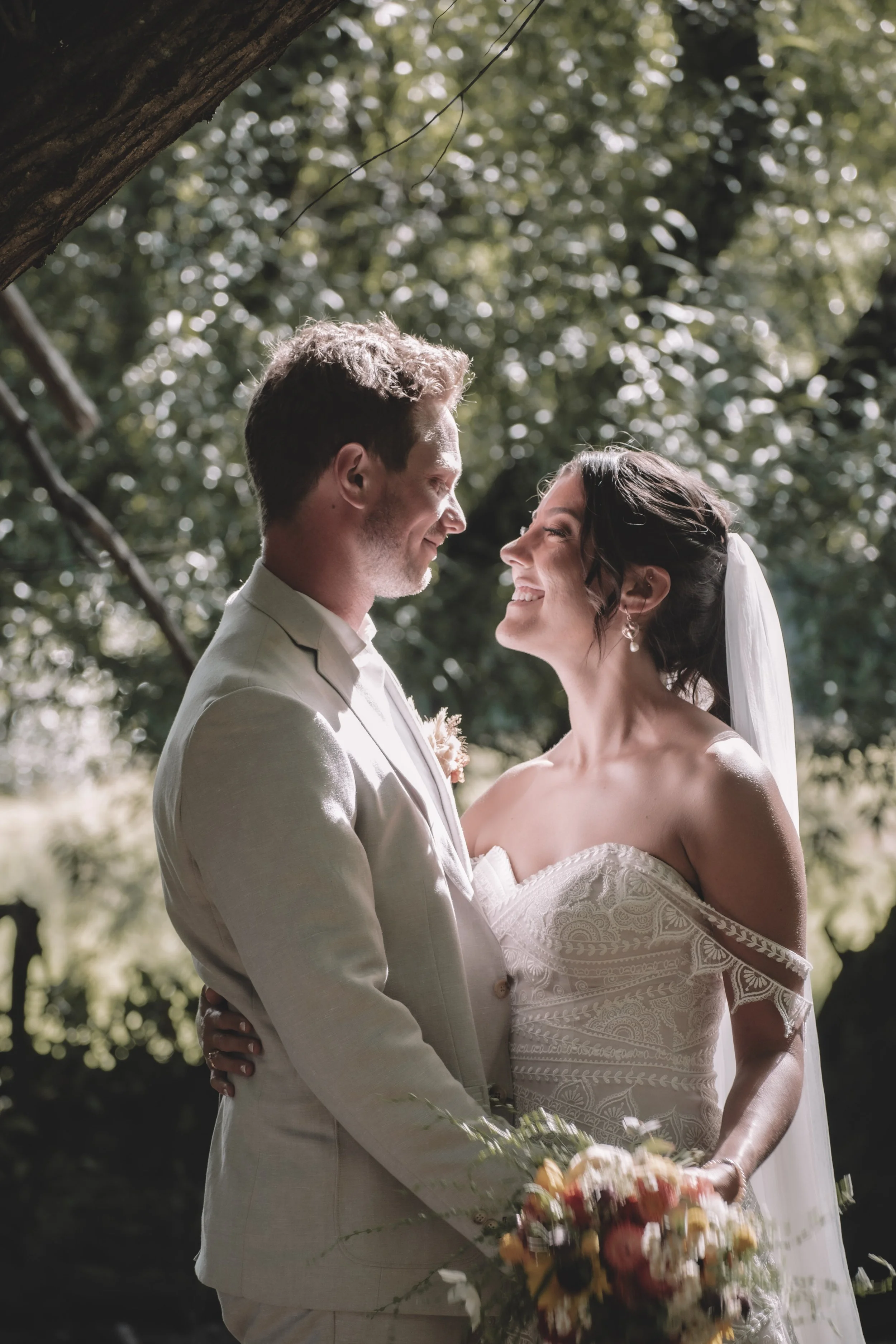 A bride and groom smiling at each other outdoors, surrounded by trees, during their wedding ceremony.