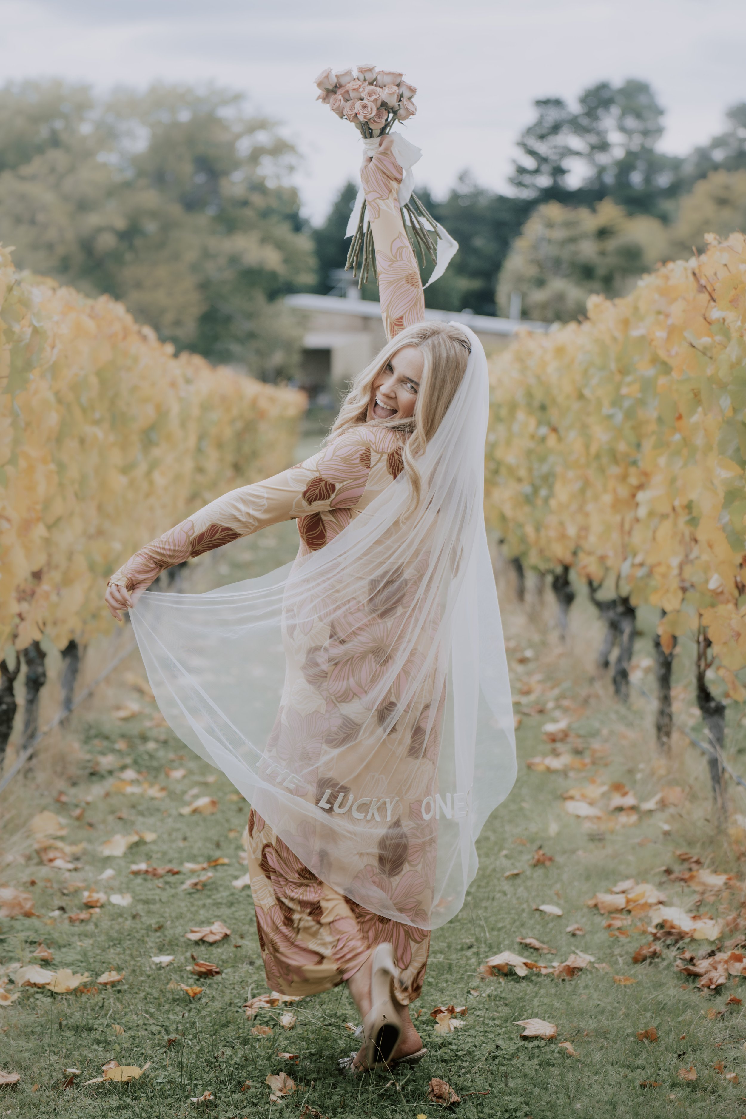 A woman in a floral dress and veil, holding a bouquet of pink roses, in a vineyard with yellow autumn leaves, smiling and celebrating.