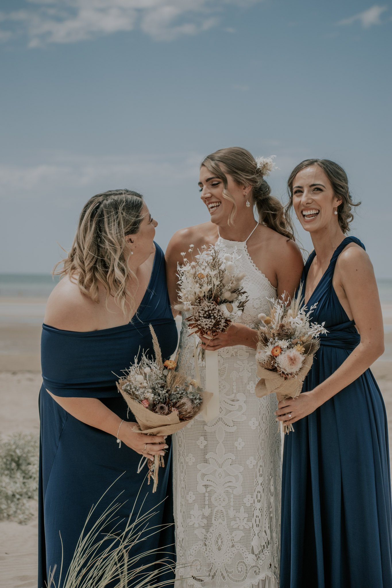 Bride and two bridesmaids on a beach, smiling and holding bouquets.