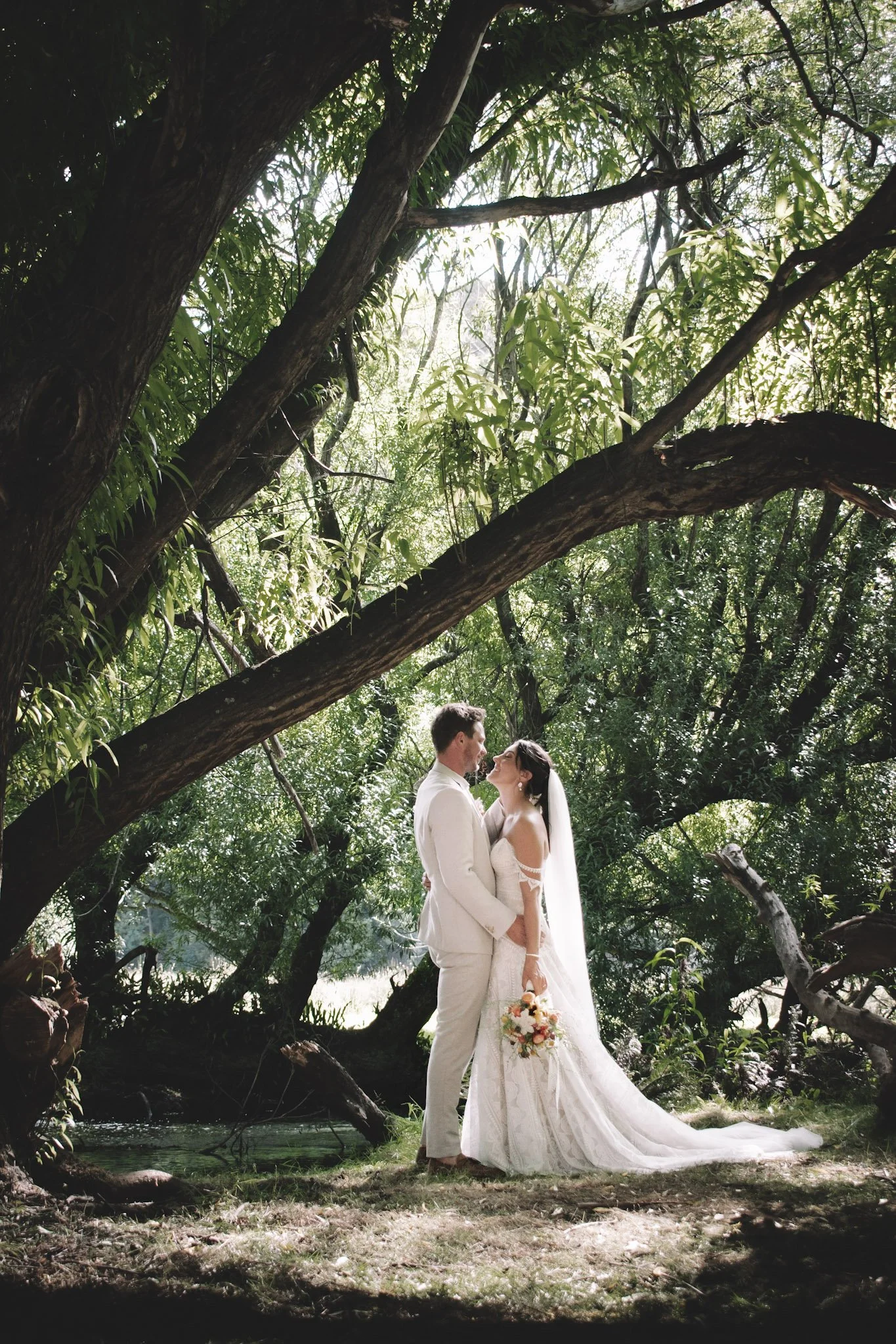 A bride and groom in wedding attire standing close together in a wooded outdoor setting, surrounded by lush green trees, with sunlight filtering through the leaves.
