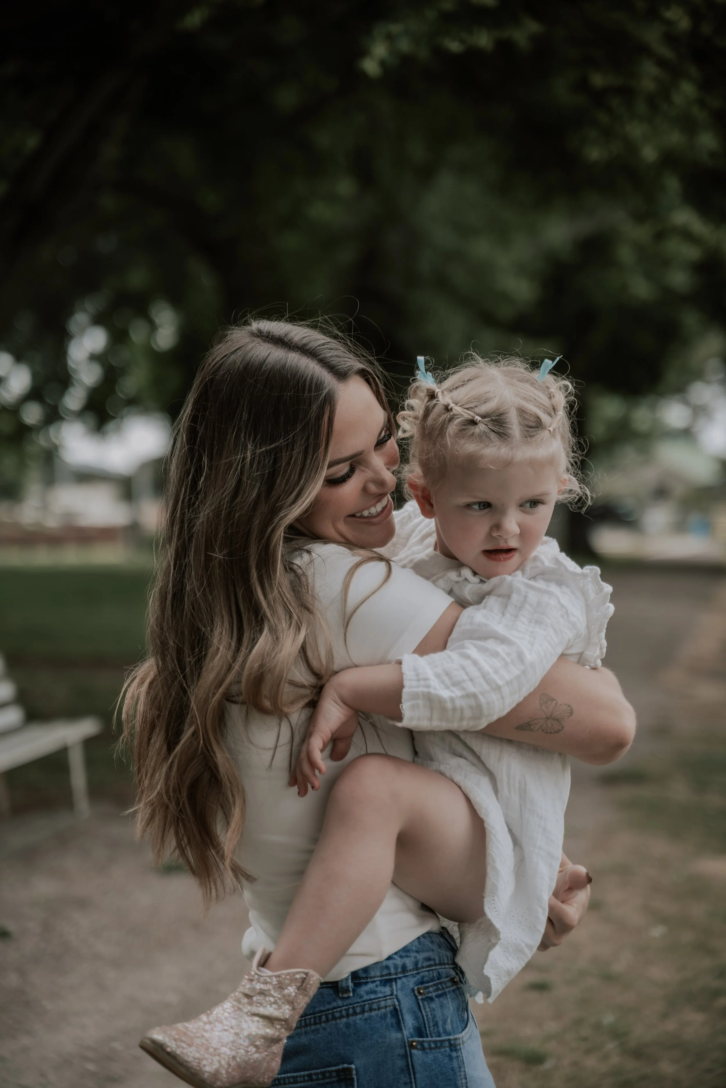 A woman with long wavy hair holding a young girl with braided hair in a park.