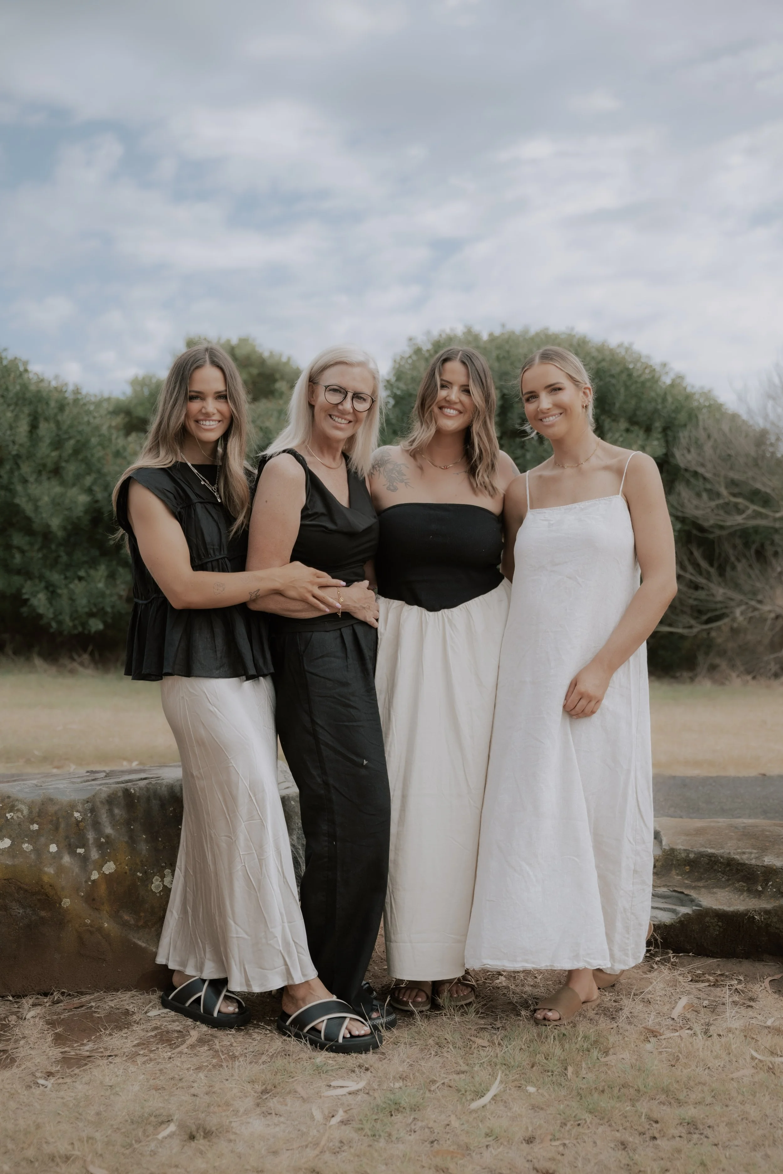 Four women standing outdoors on grass, smiling at the camera, with trees and a cloudy sky in the background.