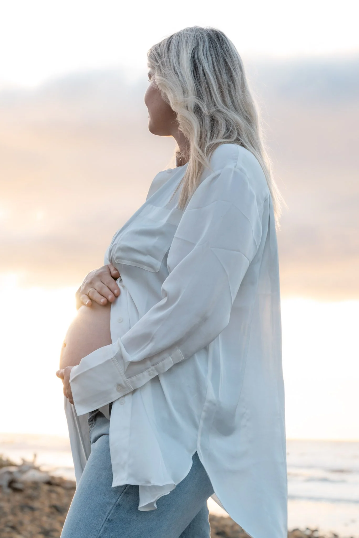 Pregnant woman with silver hair standing on a beach at sunset, holding her belly and gazing into the distance.