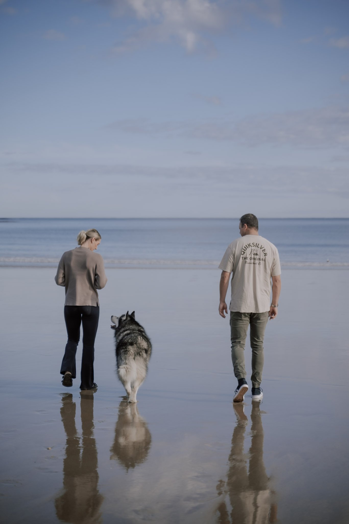 Two people walking on the beach with a husky dog, facing towards the ocean under a partly cloudy sky.
