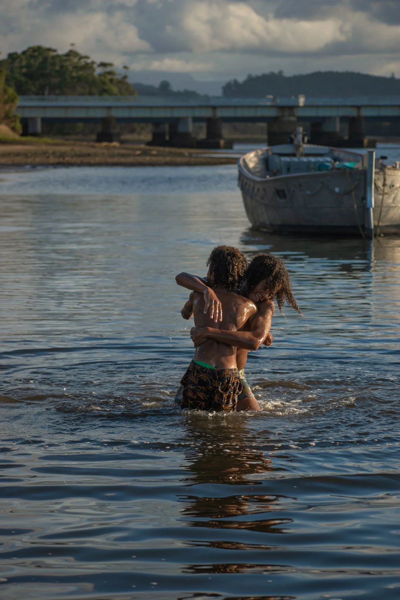 Two children embracing in the water near a boat, with a bridge and landscape in the background during sunset.