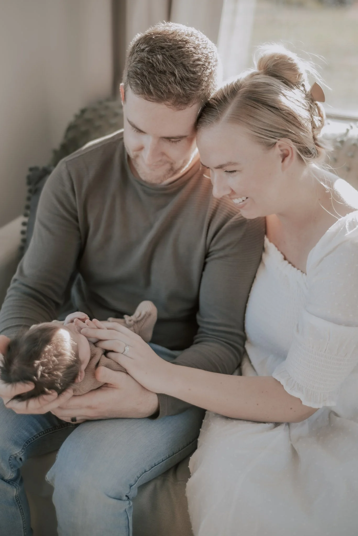A couple sitting together holding a newborn baby, smiling and looking at the baby, in a cozy indoor setting.