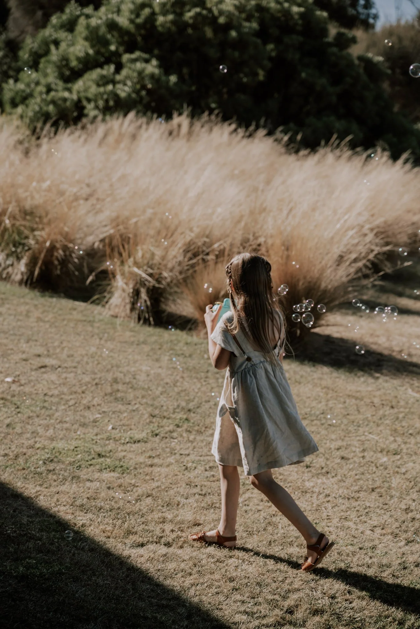 A young girl with long brown hair playing with bubbles outdoors, surrounded by tall grass and trees, during daytime.