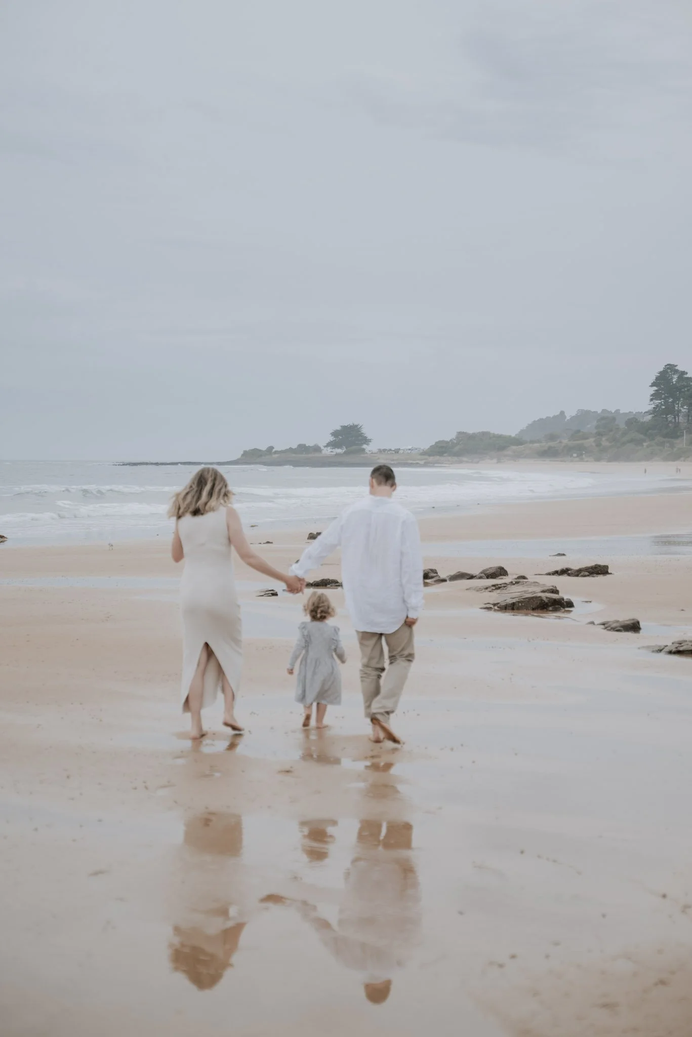 A family of three walking hand-in-hand on a beach, with the ocean, rocks, and distant trees in the background during cloudy weather.
