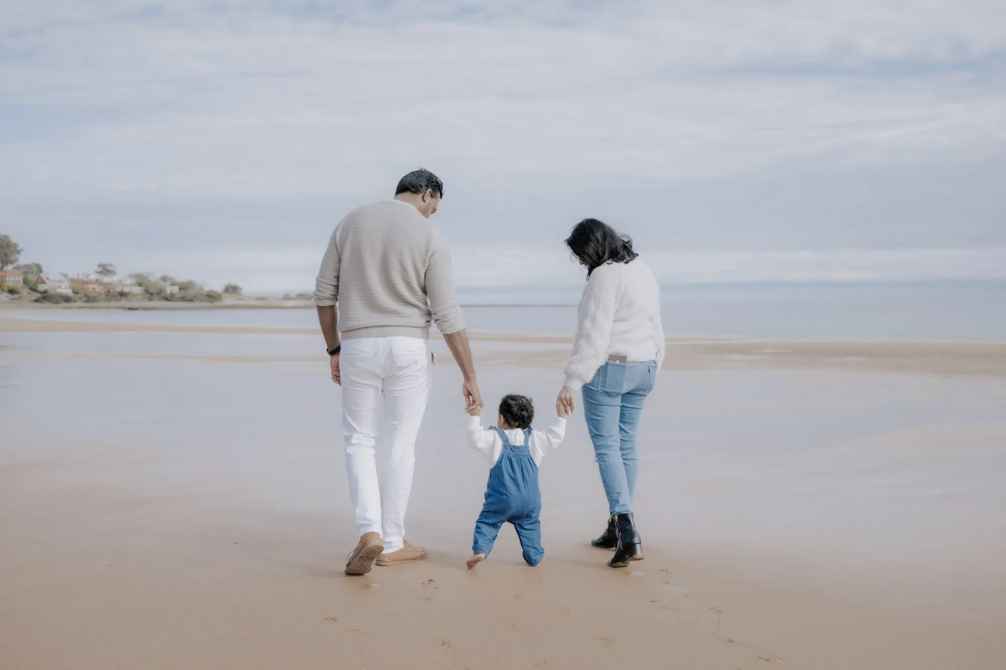 A family of three, a man, a woman, and a young girl, walking on the beach holding hands with shallow tide water and houses in the background.