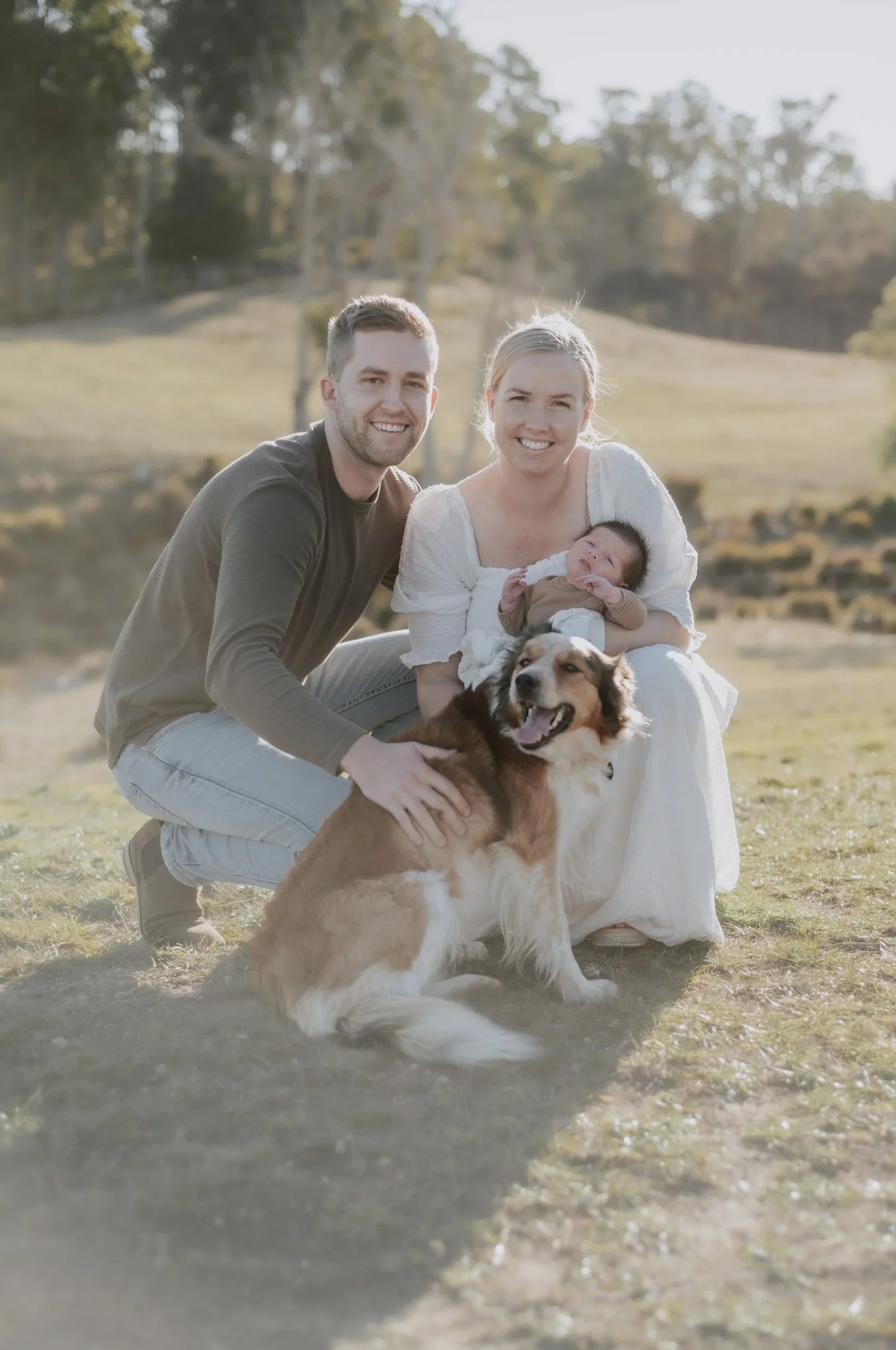 A happy family with a man, woman, baby, and dog outdoors in a grassy field with trees in the background, smiling at the camera.