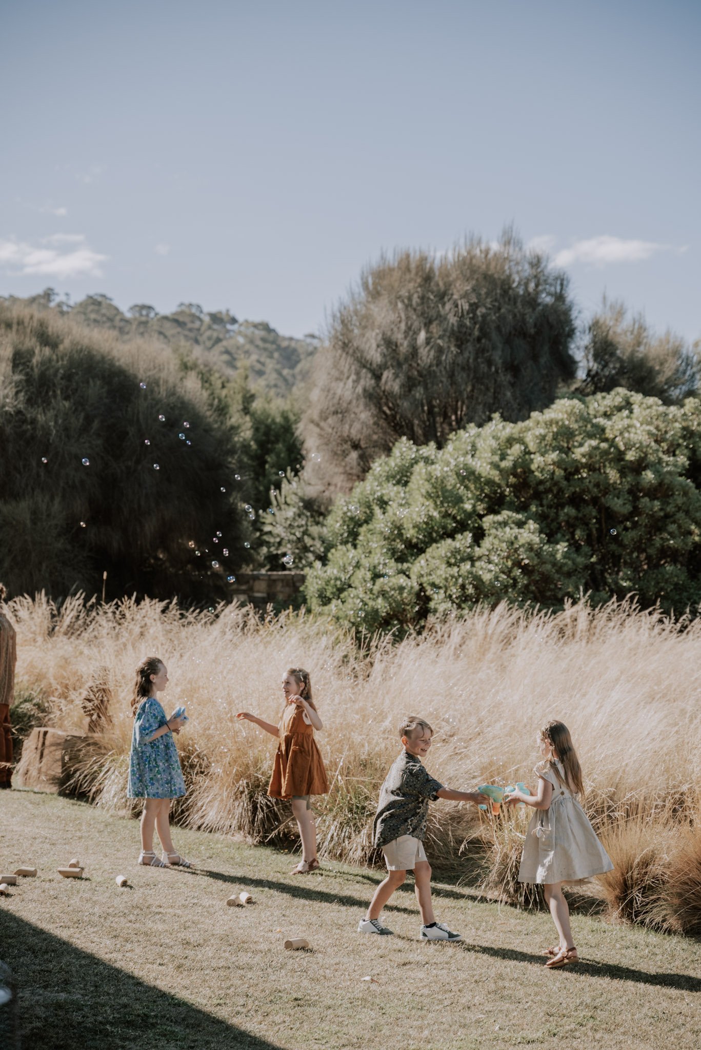 Four young children in colorful dresses playing outside, blowing bubbles and holding cups with water, on a grassy yard with tall dried grass and lush green trees in the background under a clear blue sky.