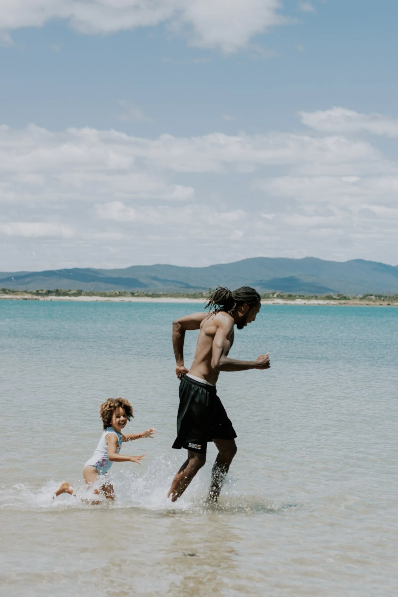 A man and a young girl running in shallow water at the beach with mountains in the background and a partly cloudy sky.