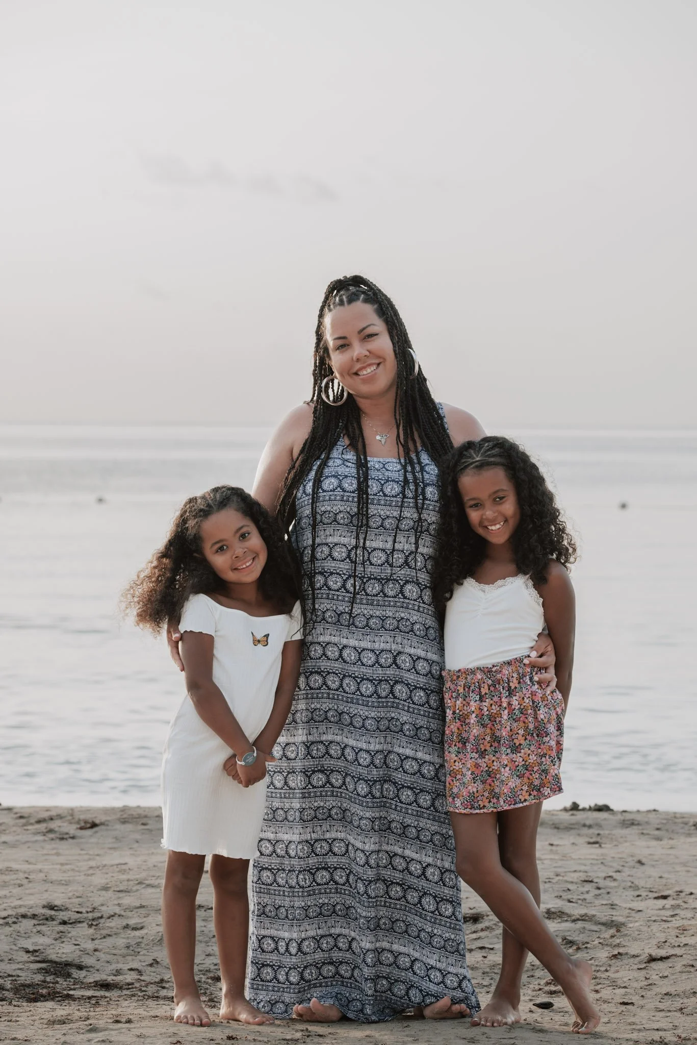A woman with two young girls stands on a beach near the ocean during sunset, smiling at the camera.