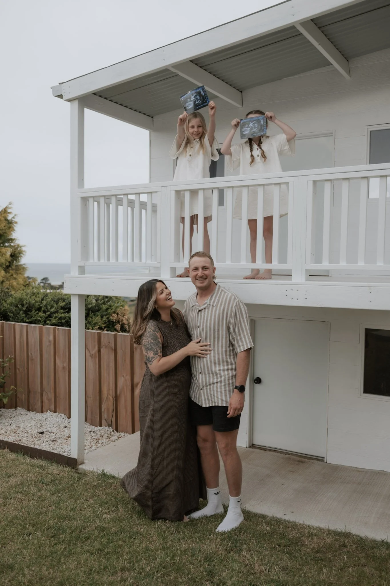 A happy couple standing outside in front of a white house, smiling and embracing each other, with two young girls on the balcony above holding ultrasound images in front of their faces.