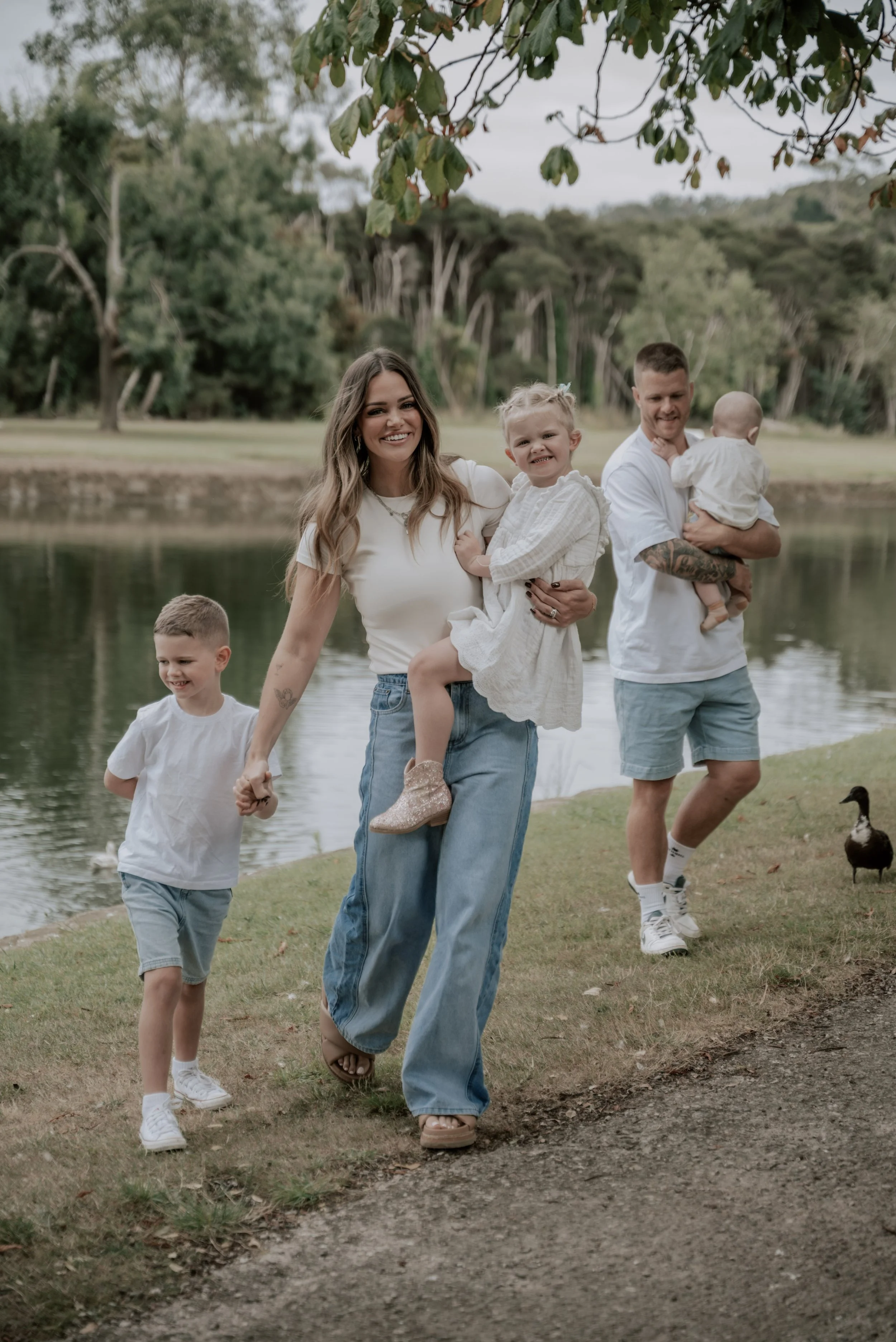 A happy family of five, including a woman, two young boys, and two young girls, walking near a lake in a park with trees in the background. The woman is holding one girl, the boy is holding the woman's hand, and a man is carrying a baby while walking beside them.