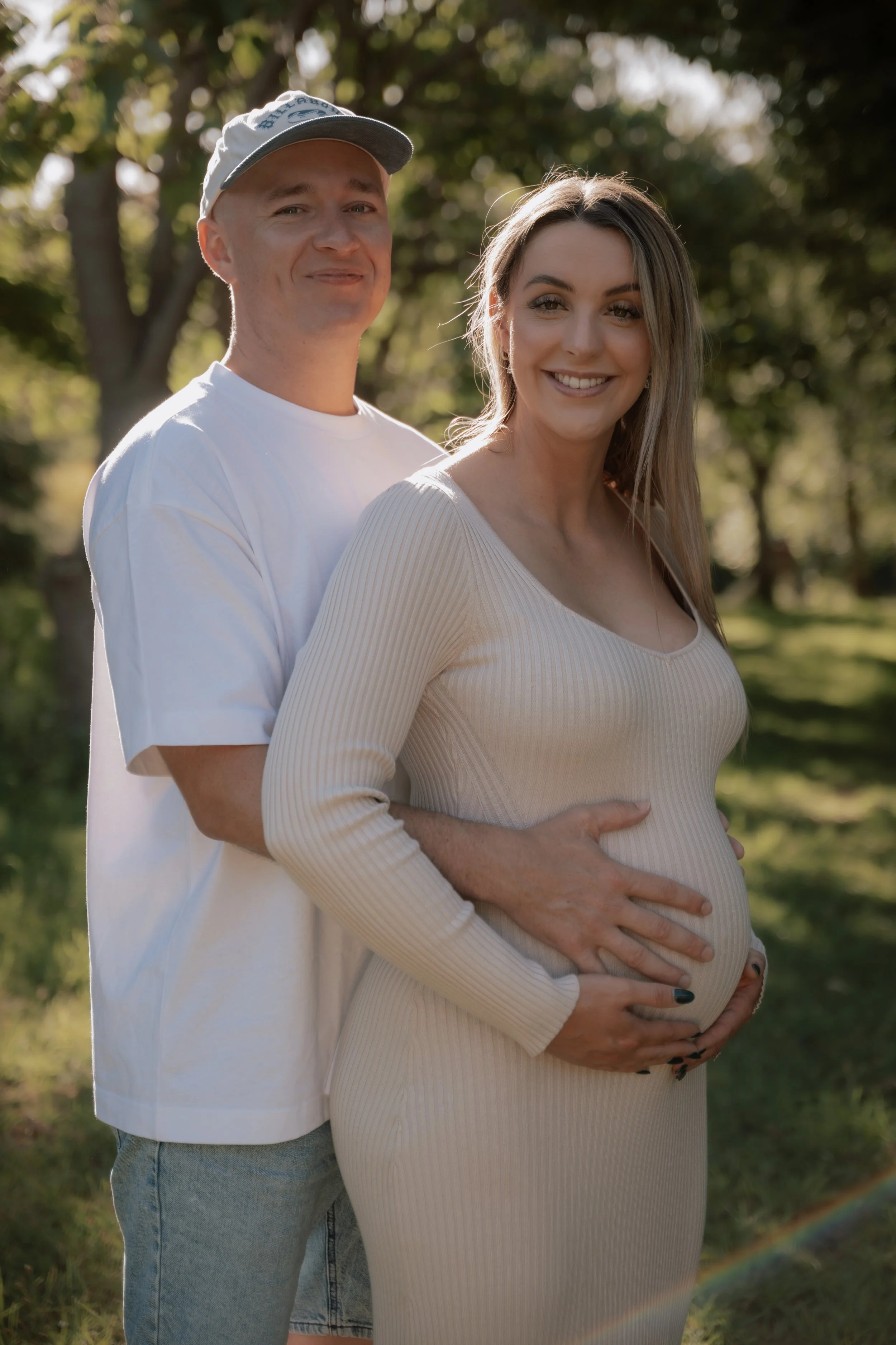 A pregnant woman with long, brunette hair smiling, being embraced from behind by a man wearing a baseball cap, as they stand outdoors in a park with trees in the background during daytime.