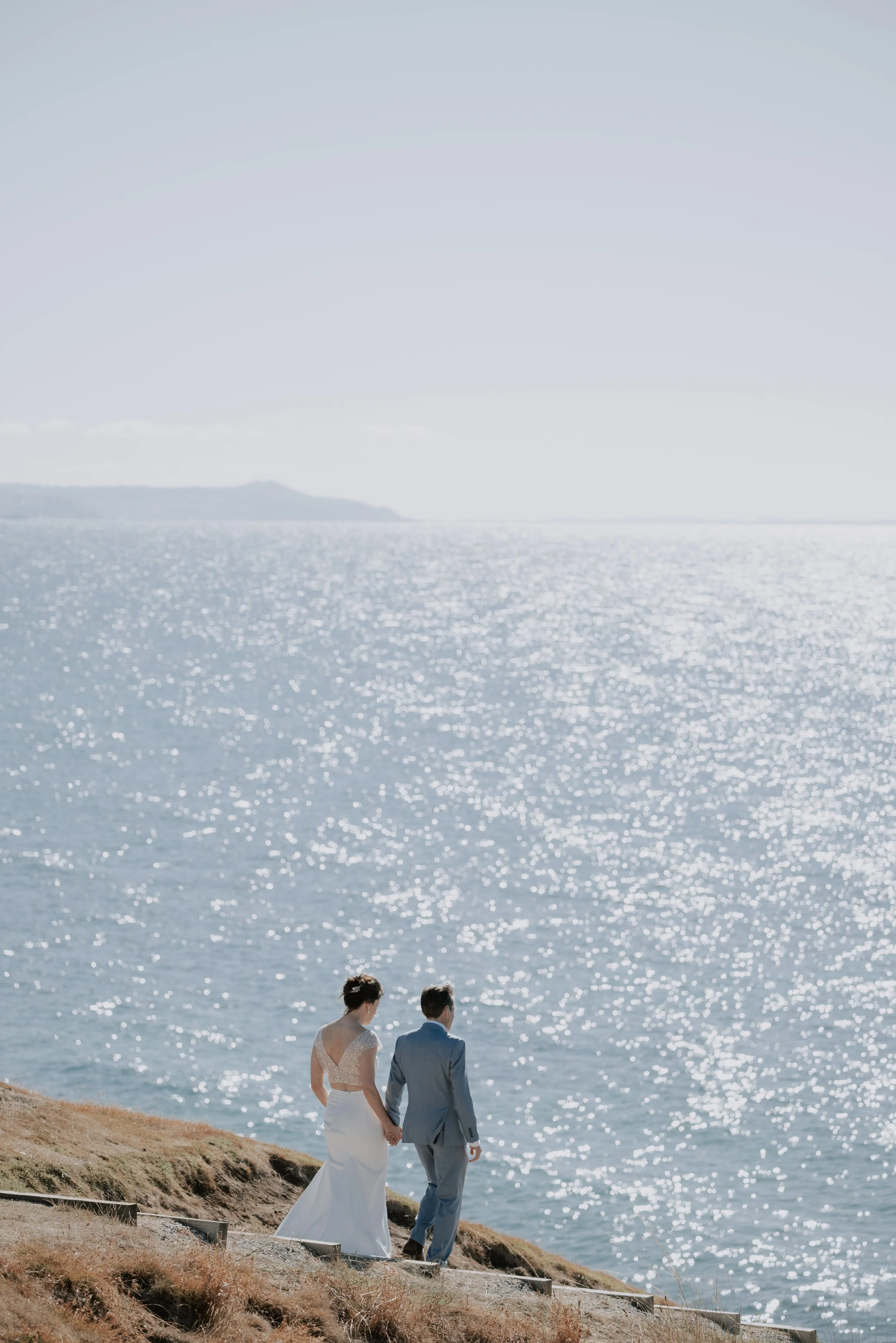 A bride and groom in wedding attire walk down the steps towards the vast sparkling ocean with mountains in the distance
