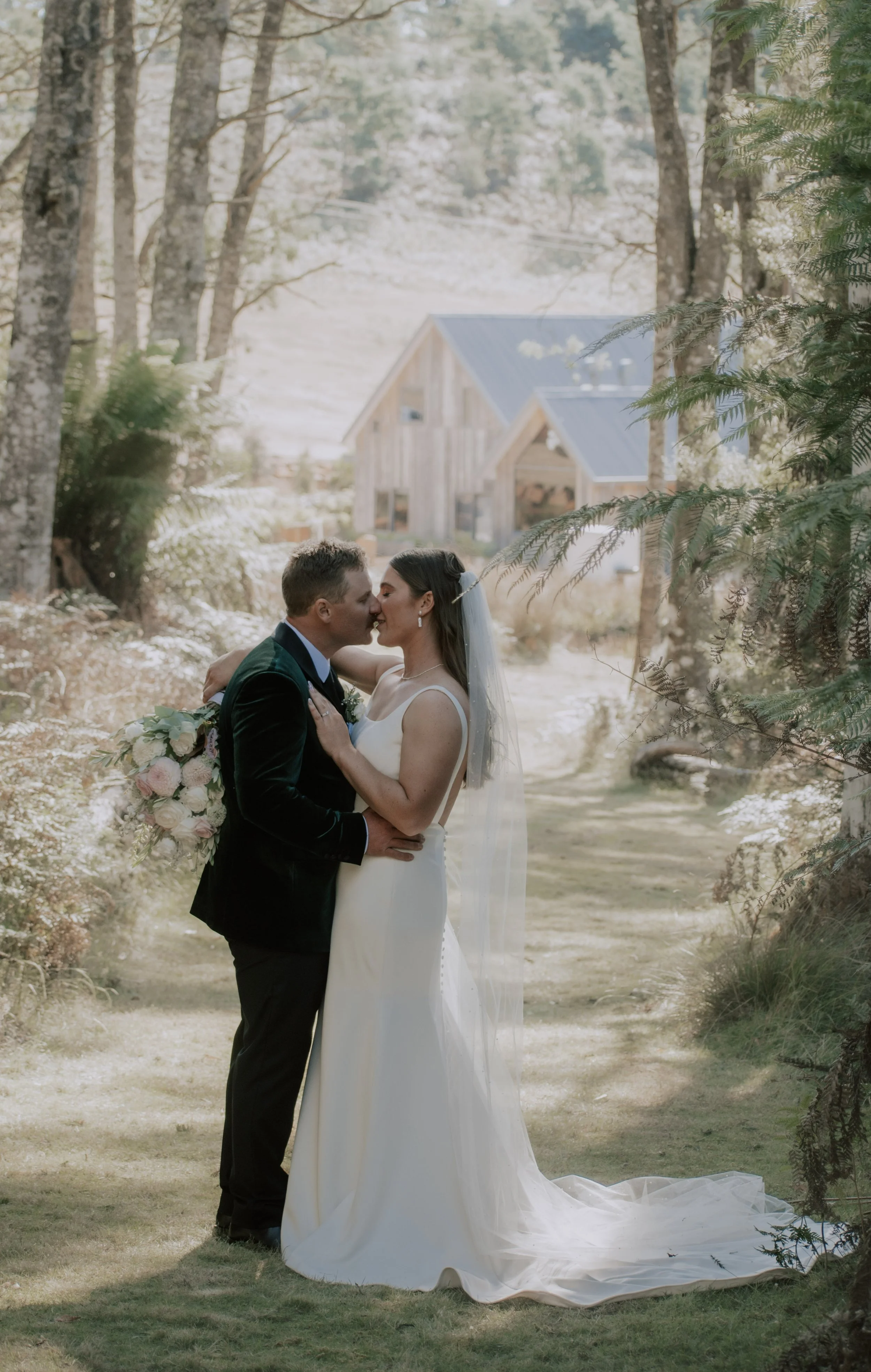 A bride and groom in wedding attire kiss in a wooded outdoor setting, surrounded by lush green trees, with sunlight filtering through the leaves.