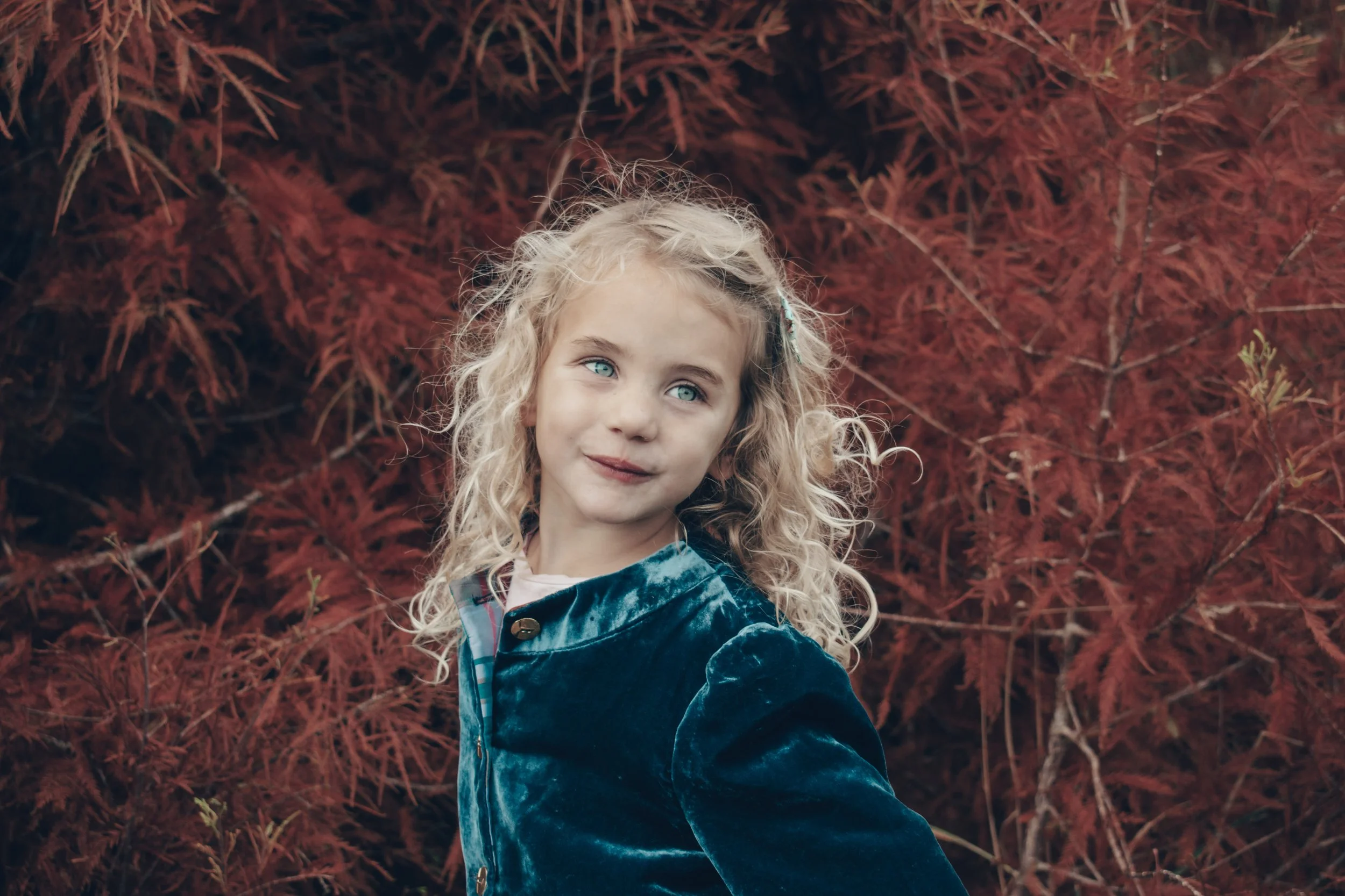 A small girl with curly blonde hair and blue eyes, wearing a blue dress smiles and chats while standing in front of red tree with autumn leaves