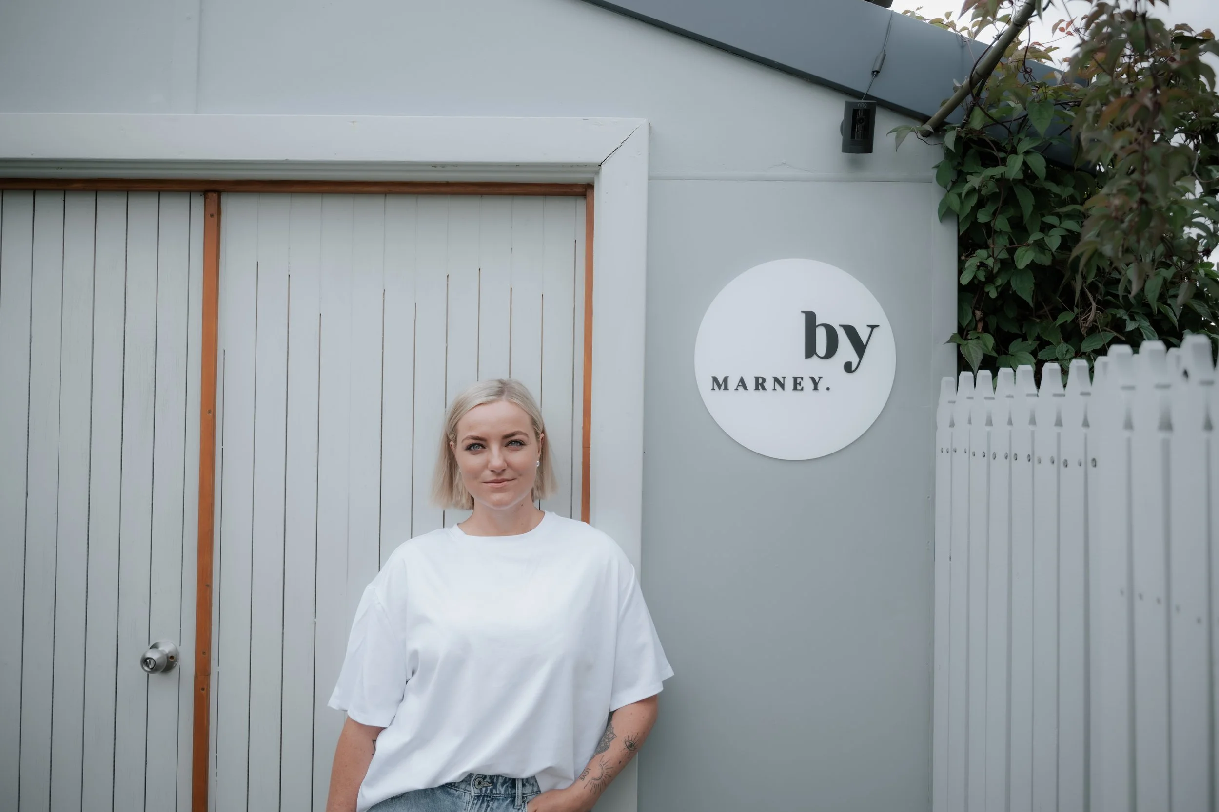 A woman with short blonde hair, wearing a white t-shirt and jeans, standing in front of a white fence and wall with a sign that says 'by MARNEY.' The background includes a gray roof, door, and some green foliage.