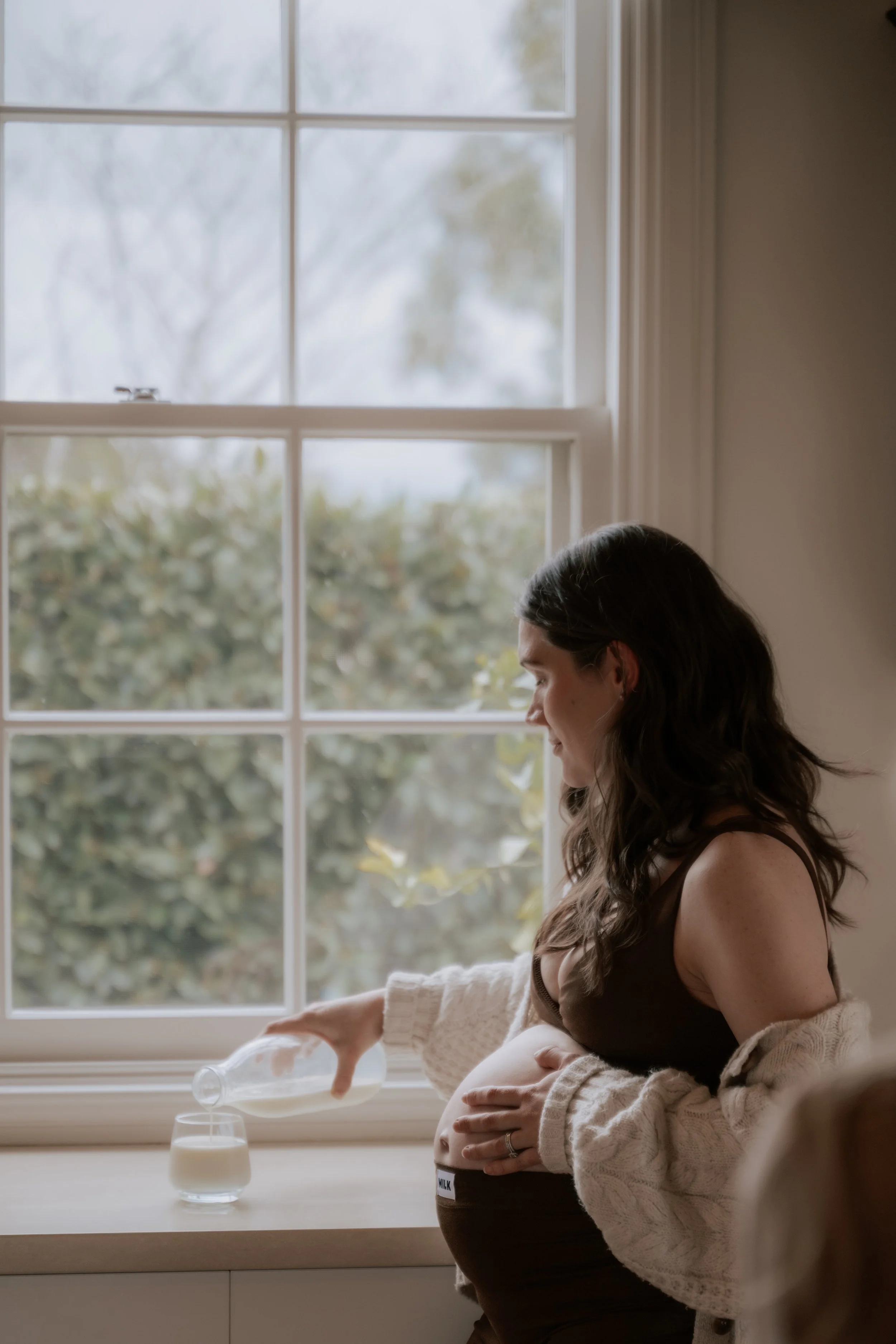 A pregnant woman wearing a black top and a cream-colored cardigan with bubble sleeves sitting by a window, pouring milk from a glass bottle into a glass. The window shows blurred outdoor trees and bushes.