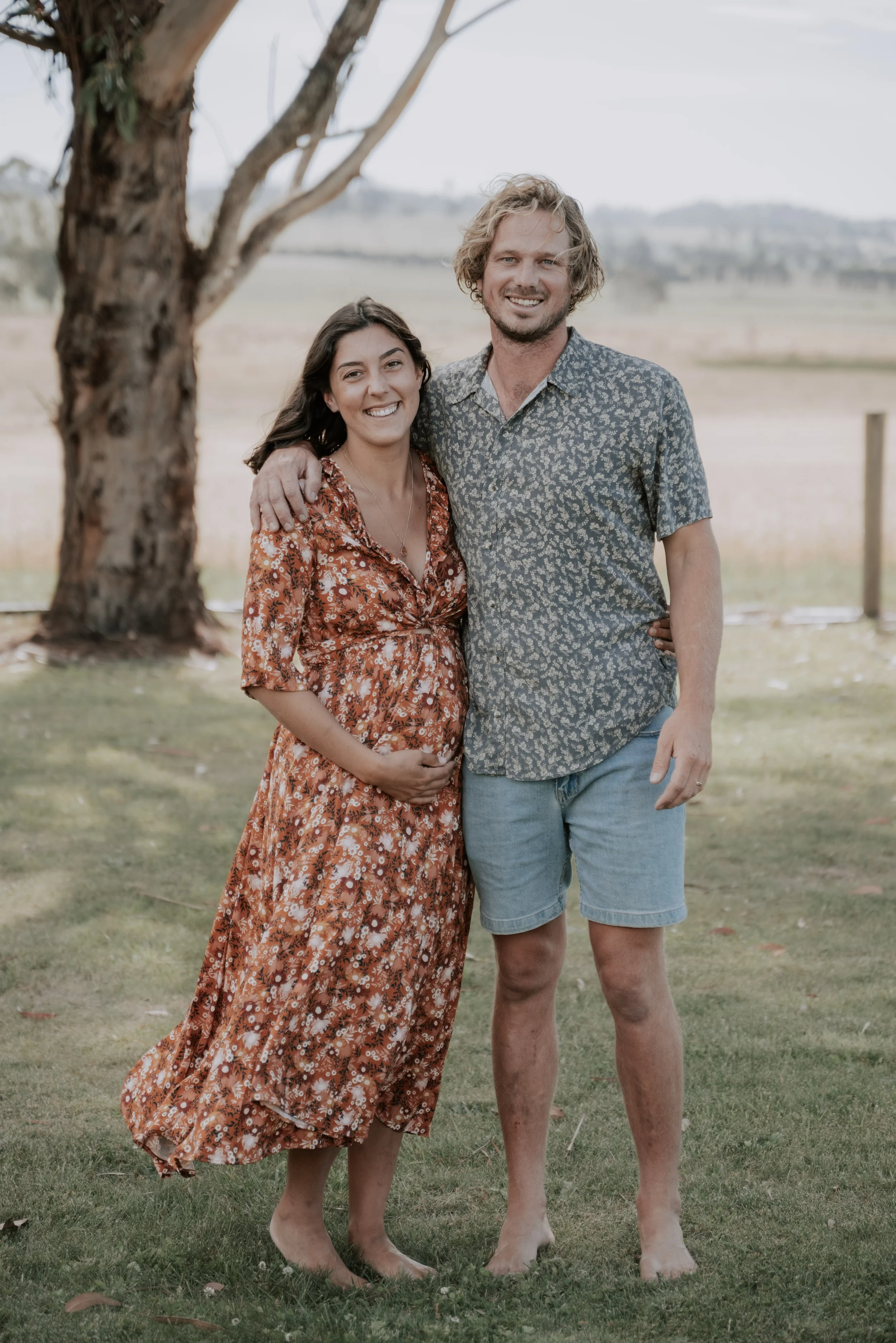 A couple smiling at the camera while outside in the afternoon with fields in the background, the woman hugging her pregnant belly.
