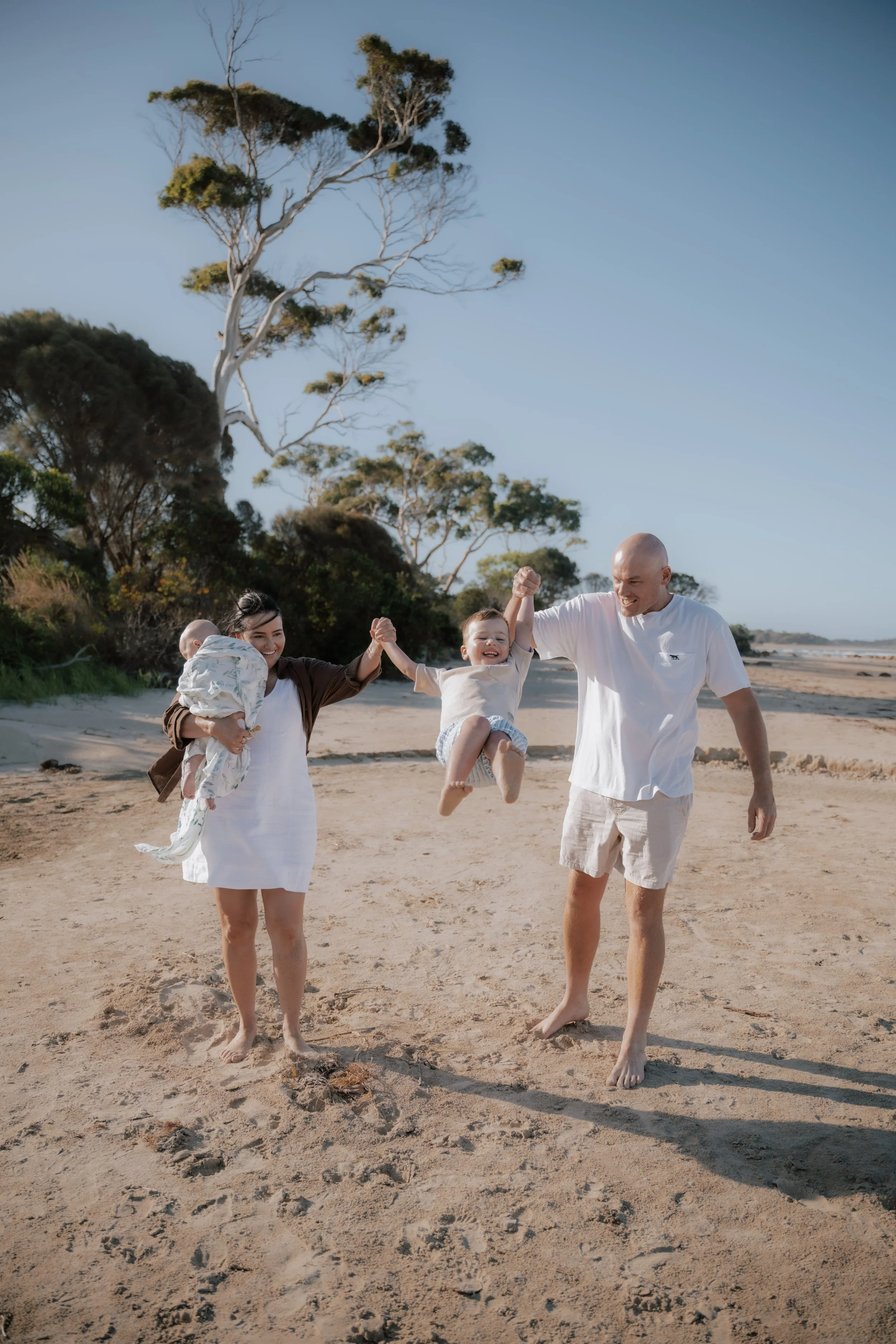 A family of four, including a man and woman holding children, enjoying a walk on a sandy beach with trees and a clear sky in the background.