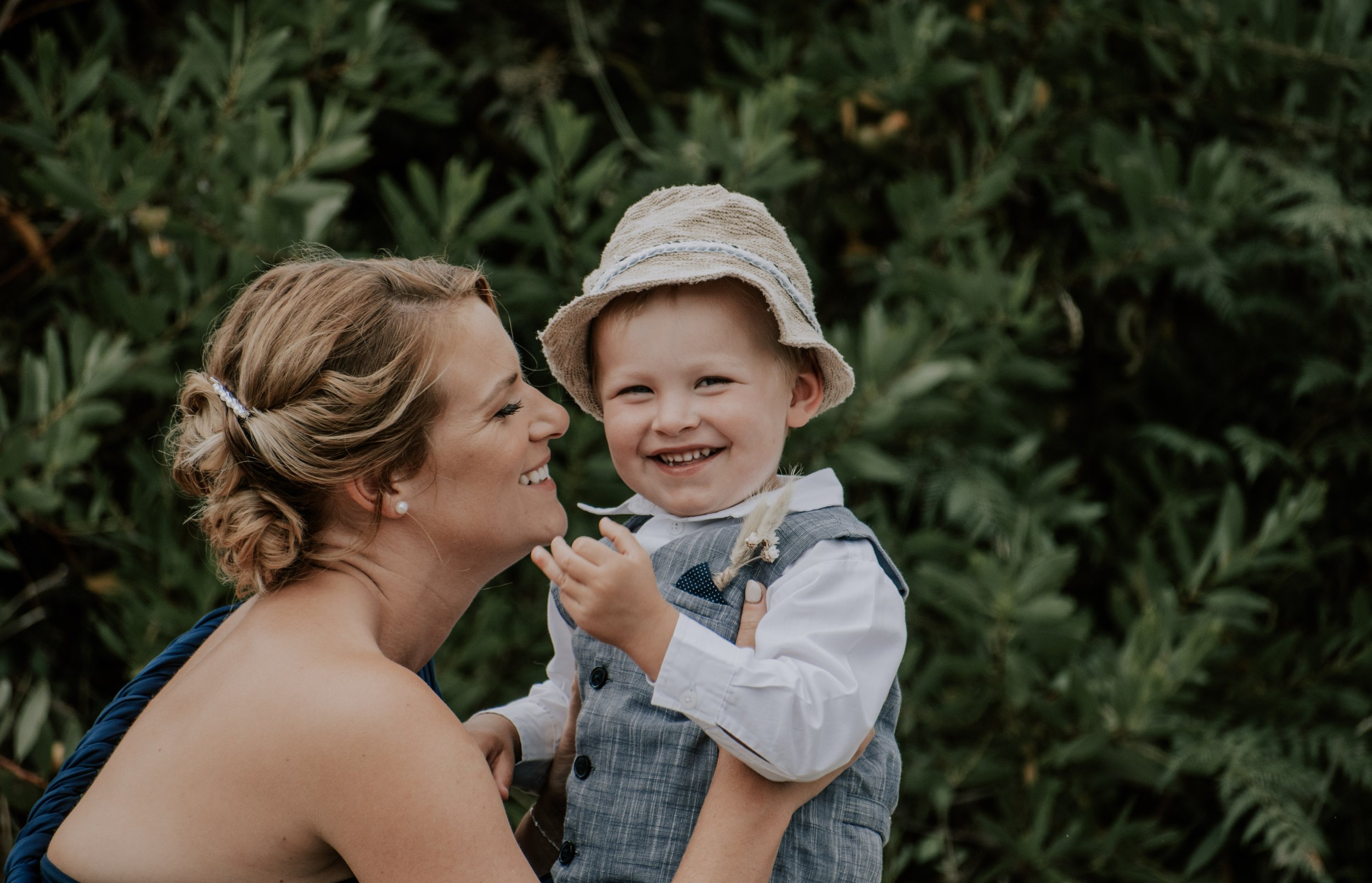 A blonde mother smiling at her son who is wearing a little suit and smiling at the camera
