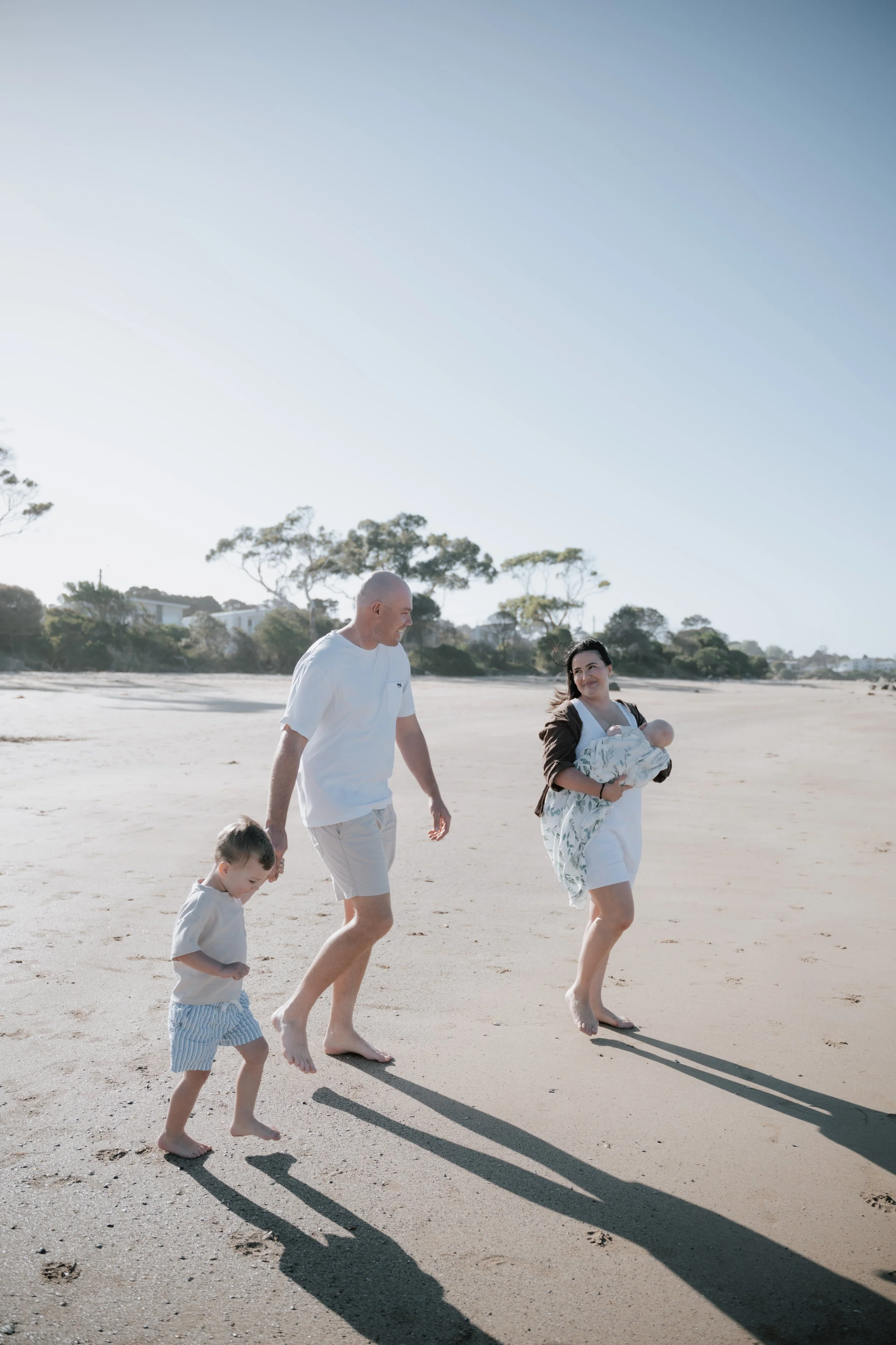 A family of four walking barefoot on a sandy beach during daytime, with the mother carrying a baby in her arms, the father holding the hand of a young boy, and shadows cast on the sand from the bright sunlight.