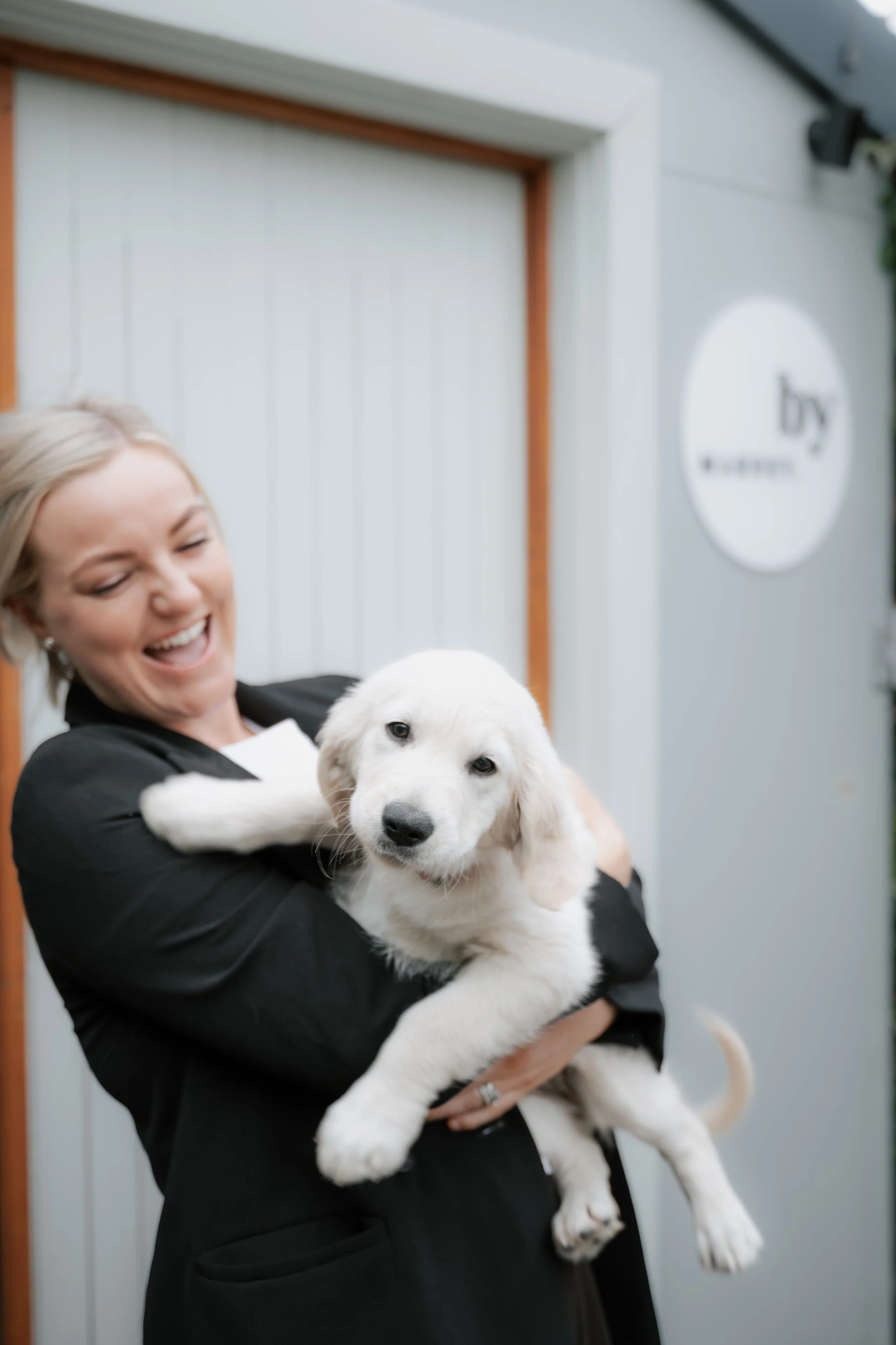 A woman smiling and holding a golden retriever puppy in her arms, standing outside near a white and gray building with a sign.