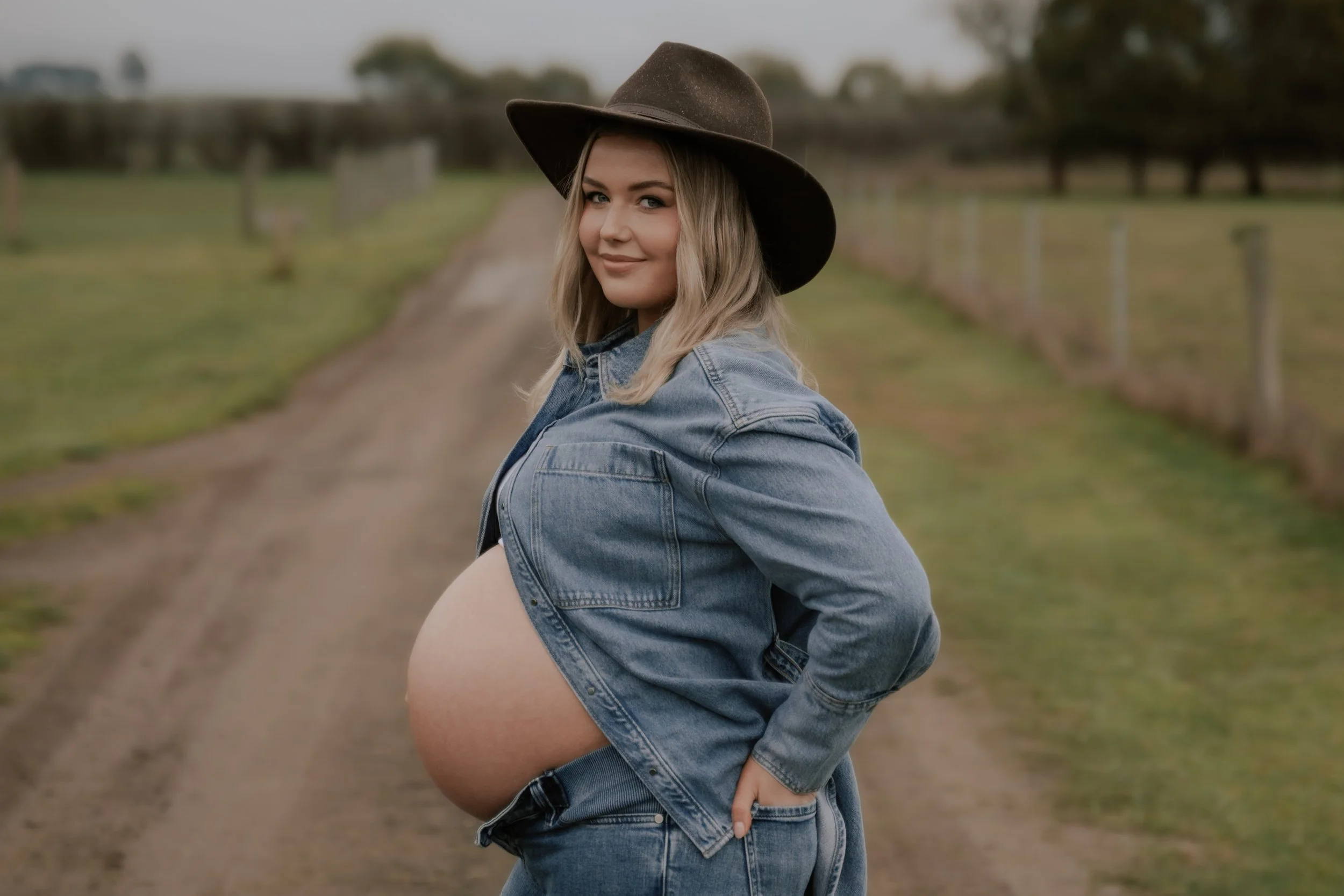 A pregnant woman with blonde hair, wearing a denim jacket and a wide-brimmed hat, sits in a field with a dirt path and trees in the background, smiling at the camera.