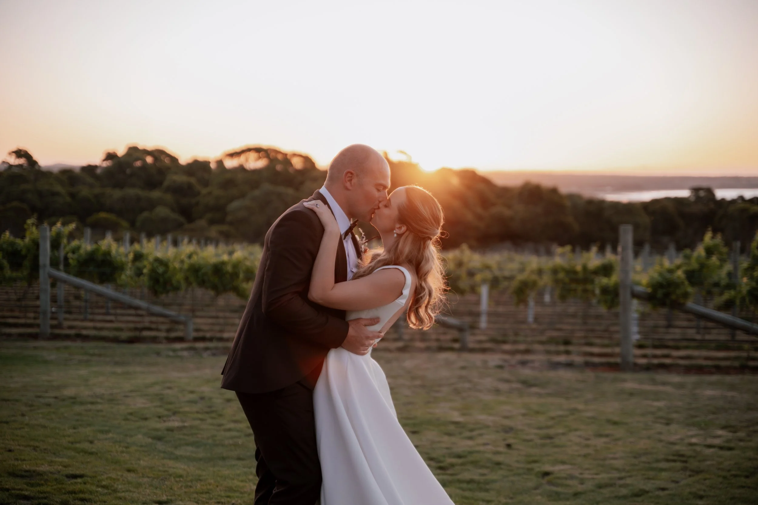 A bride and groom kiss in a vineyard during sunset, with trees and a distant horizon.