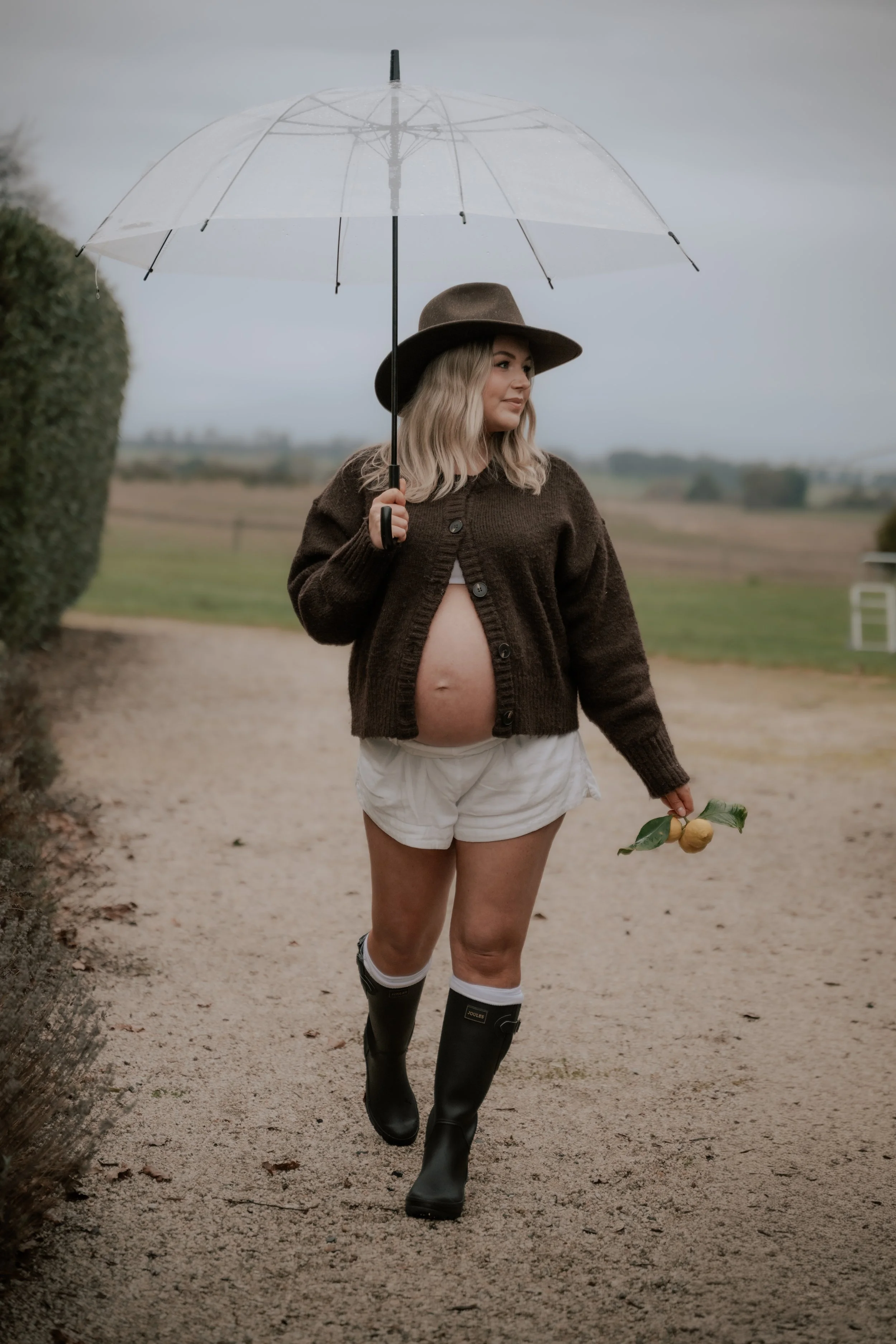 A pregnant woman walking on a dirt path outdoors on a rainy day, holding a transparent umbrella, wearing a brown cardigan, white shorts, rain boots, and a wide-brim hat, holding a yellow flower.