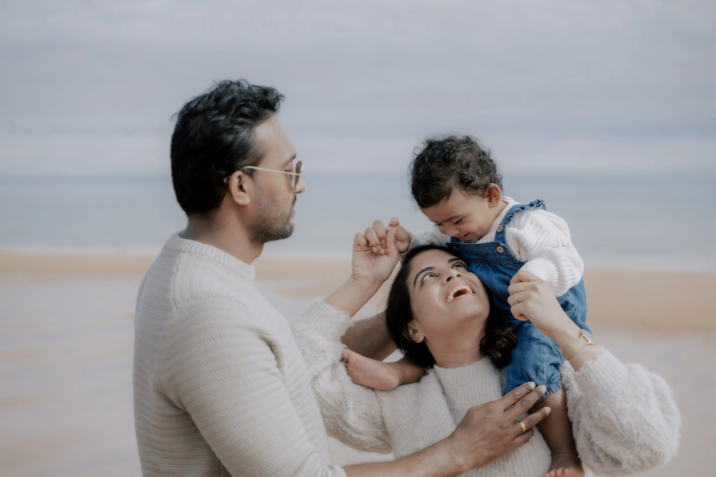 A family of three looking at each other and laughing, the baby riding her mother's shoulders at the beach 