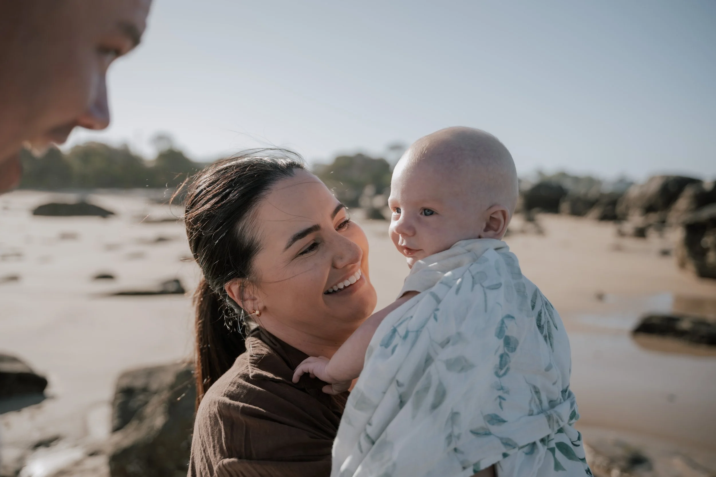 A woman is holding a smiling baby on a beach with rocks and clear sky in the background.
