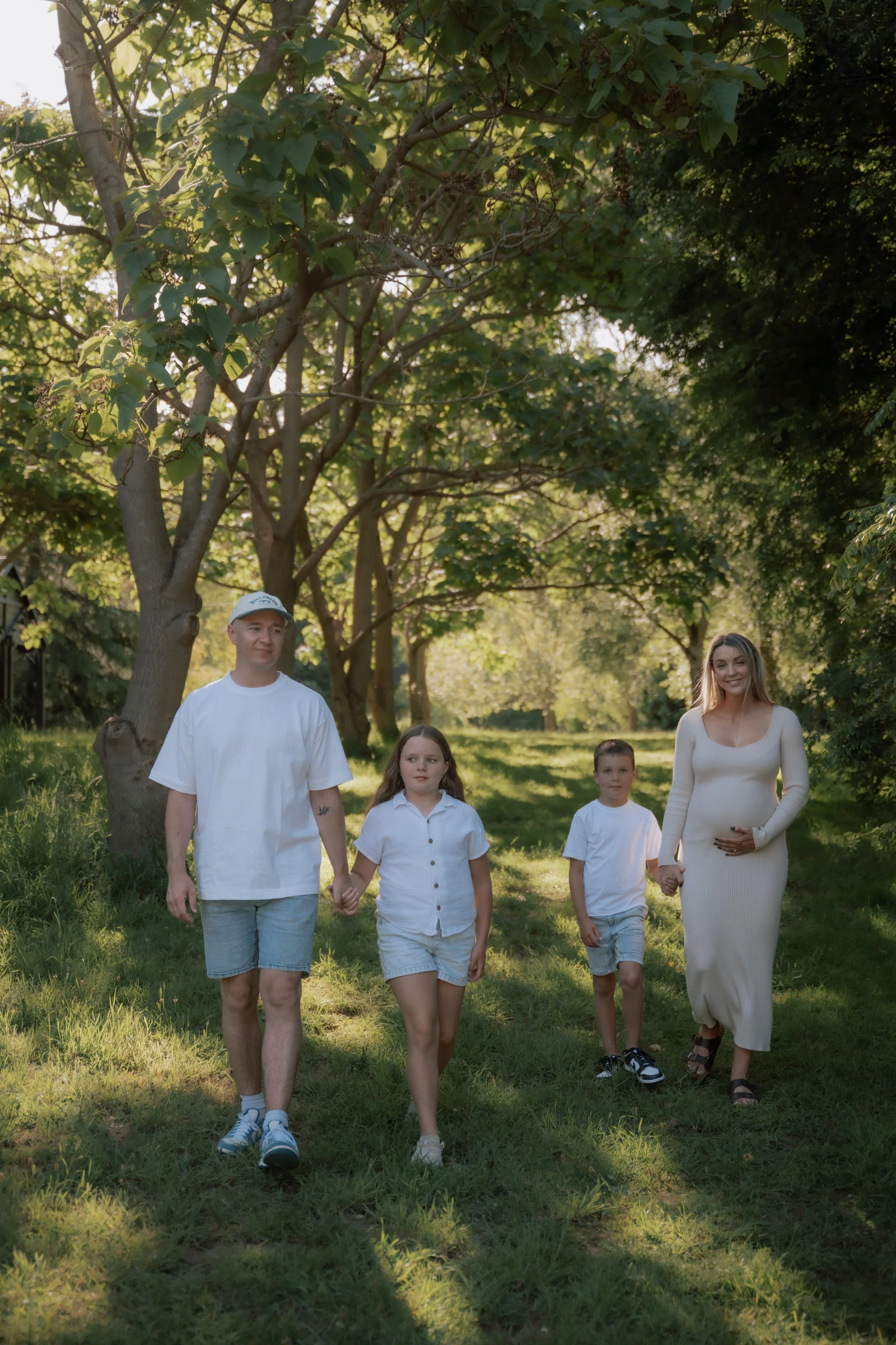 A family of five walking hand in hand through a grassy park with tall trees and sunlight filtering through the leaves.