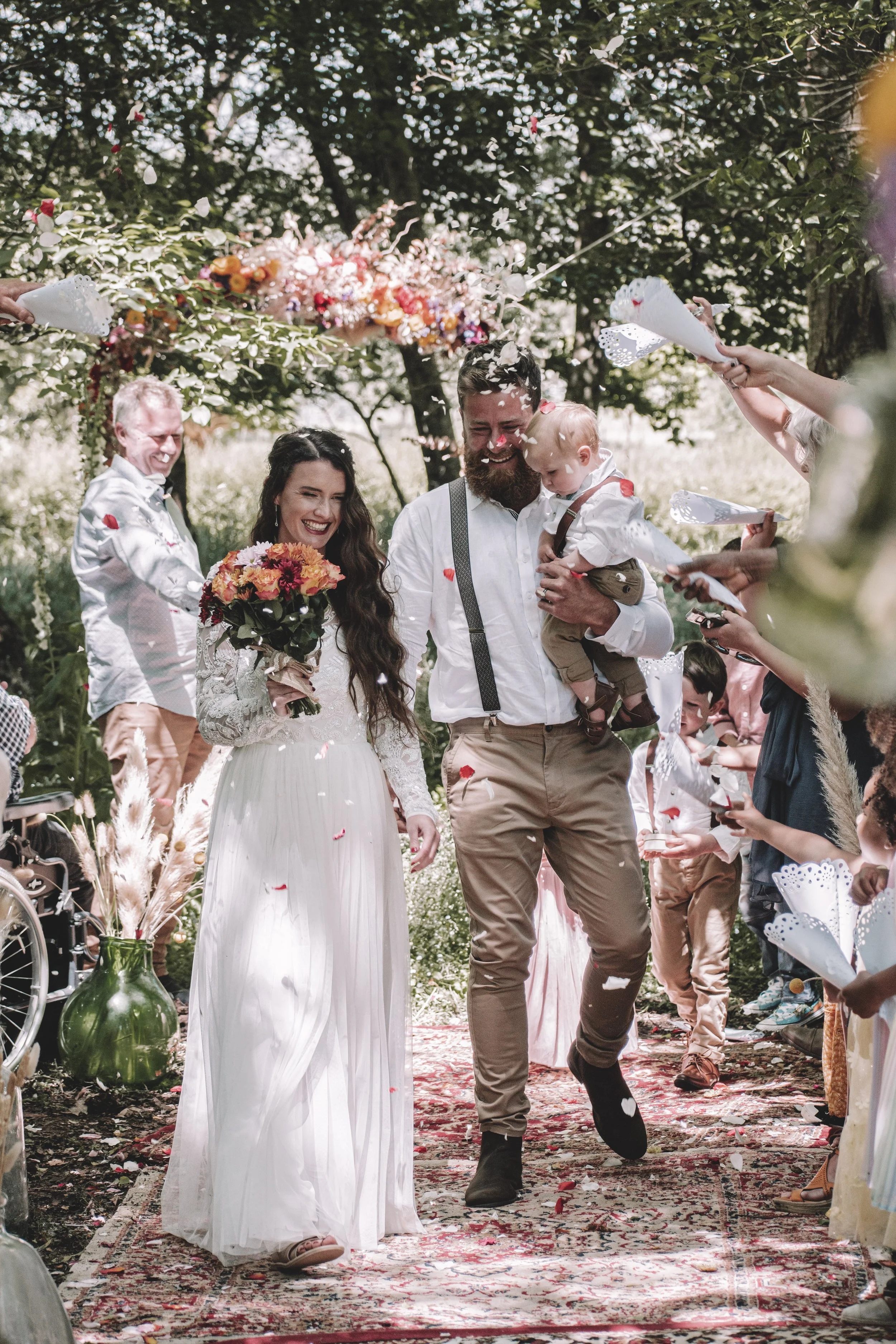 A just married couple walk down a carpeted aisle holding their baby and laughing as petals are thrown at them, in a forest setting