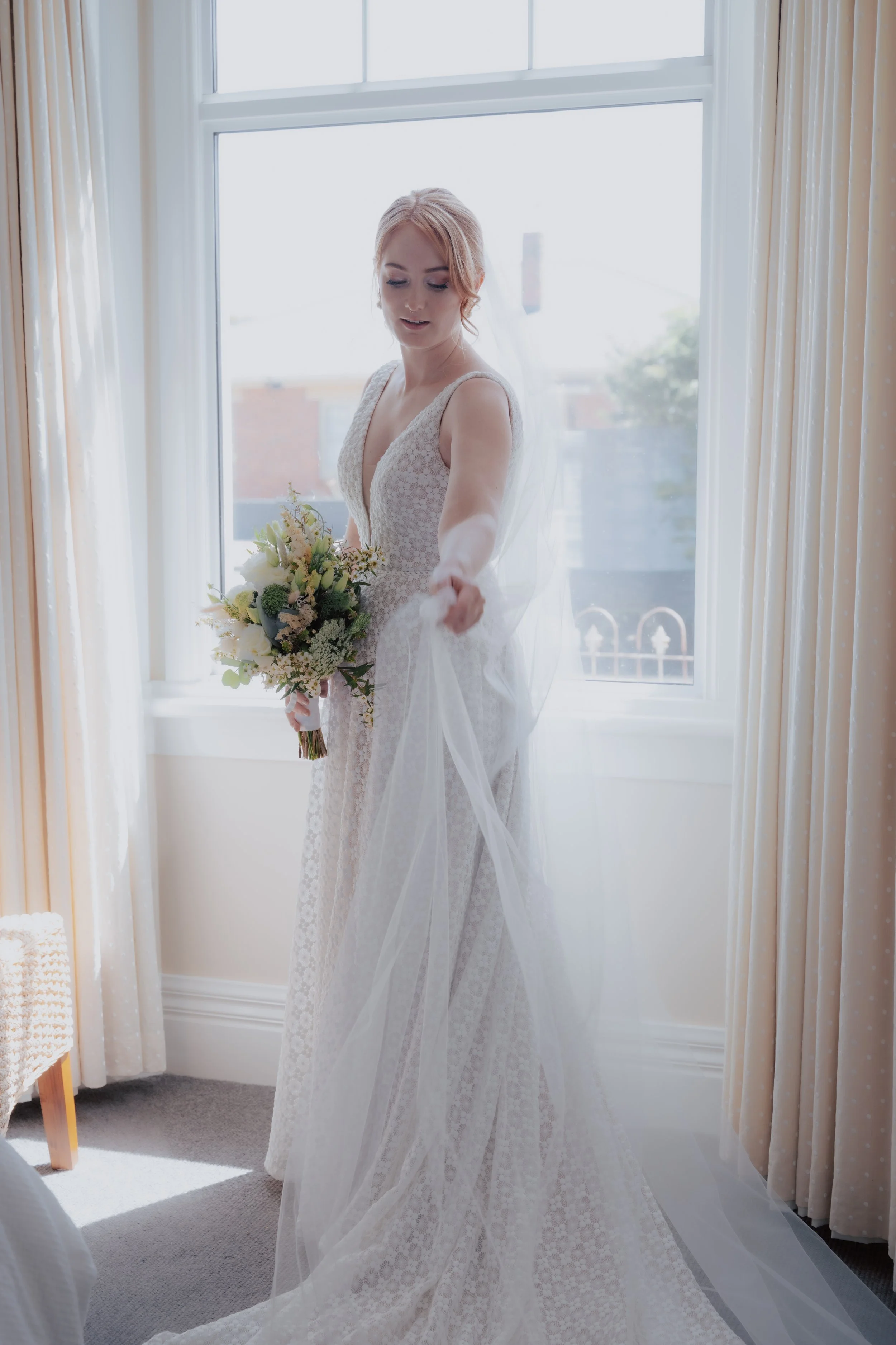A bride in a white lace wedding gown holding a bouquet of flowers, standing near a window with curtains.
