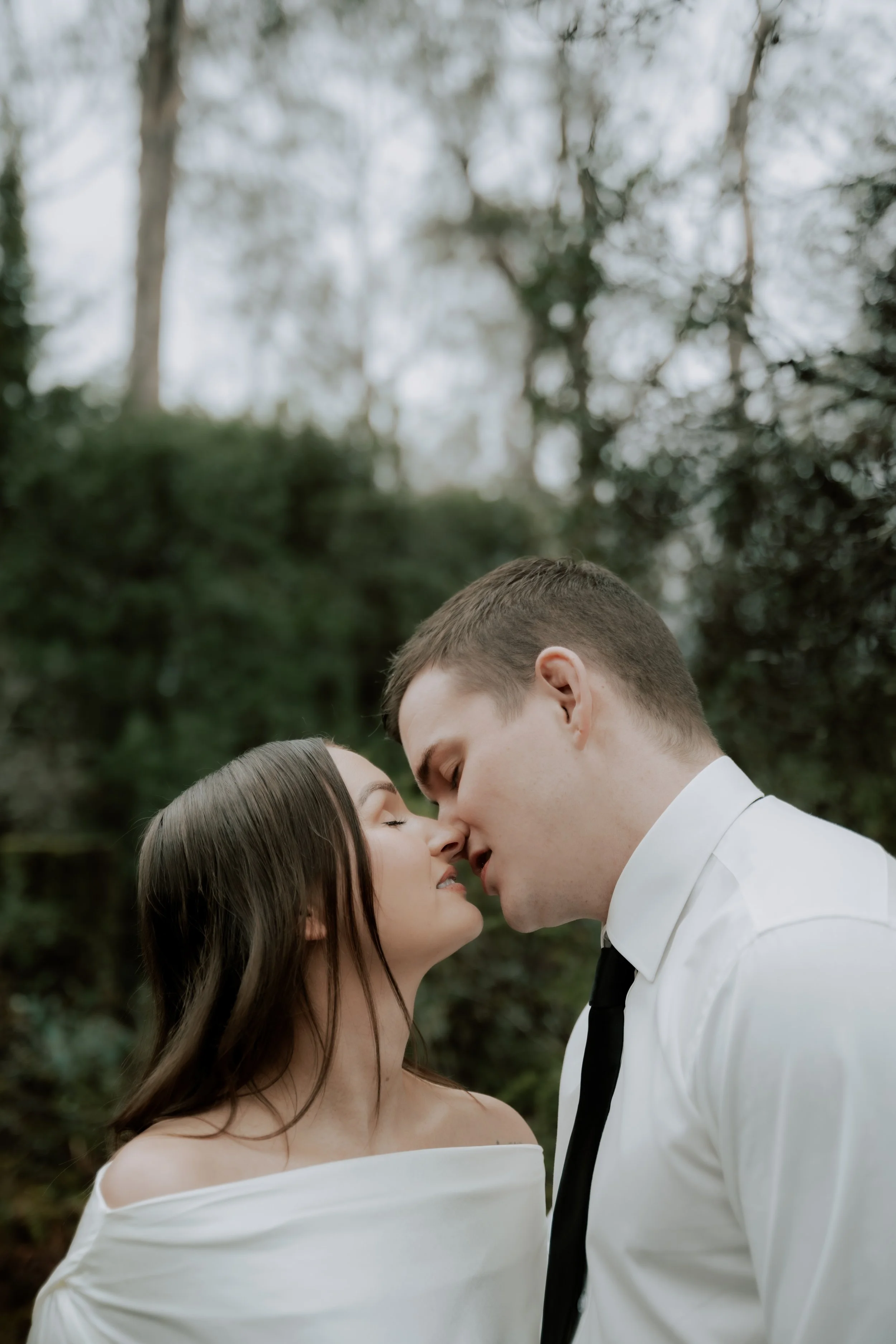 A couple is outdoors, about to kiss, with their foreheads touching, in a natural, wooded setting.