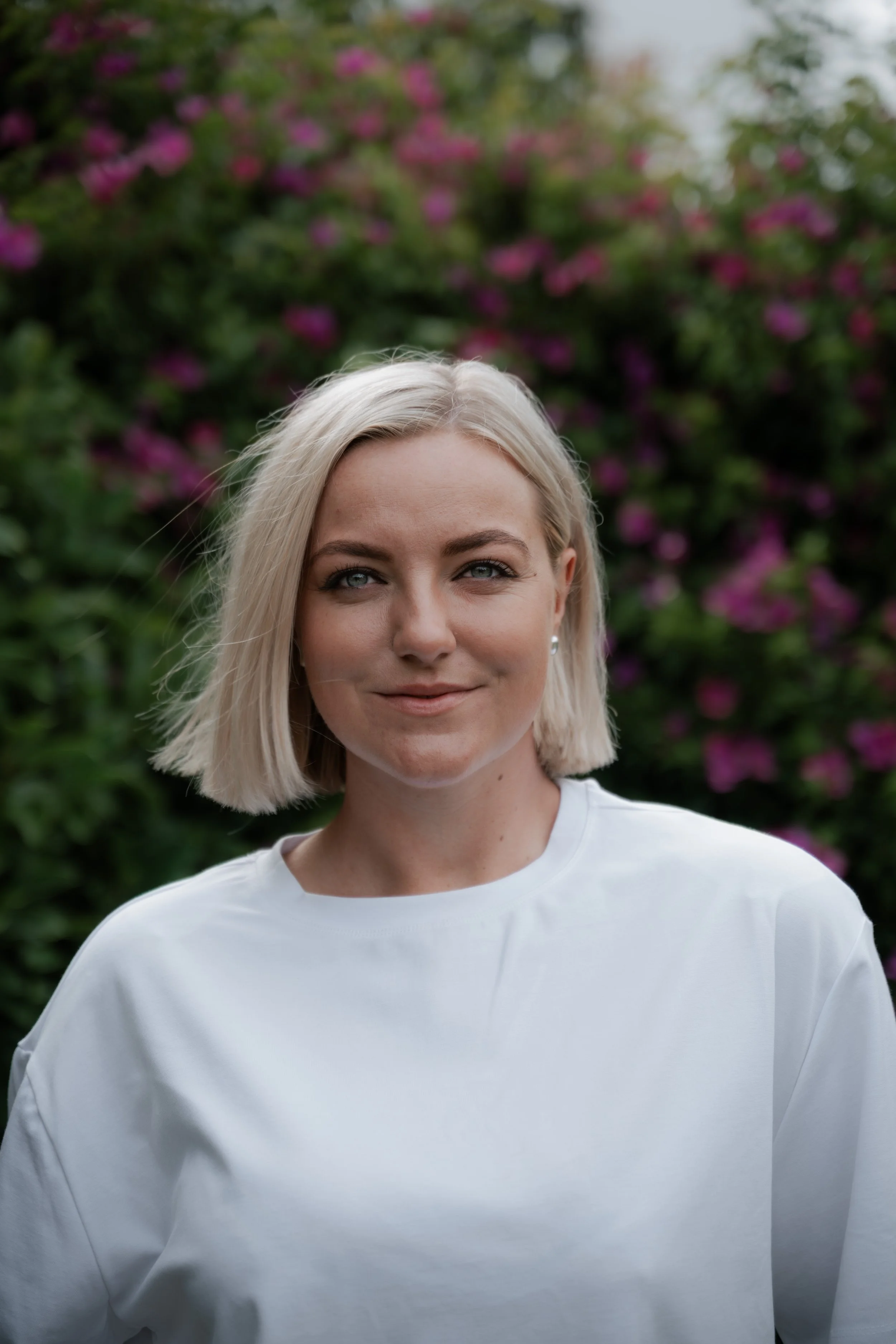 A woman with blonde hair wearing a white shirt standing outdoors in front of green foliage with purple flowers.