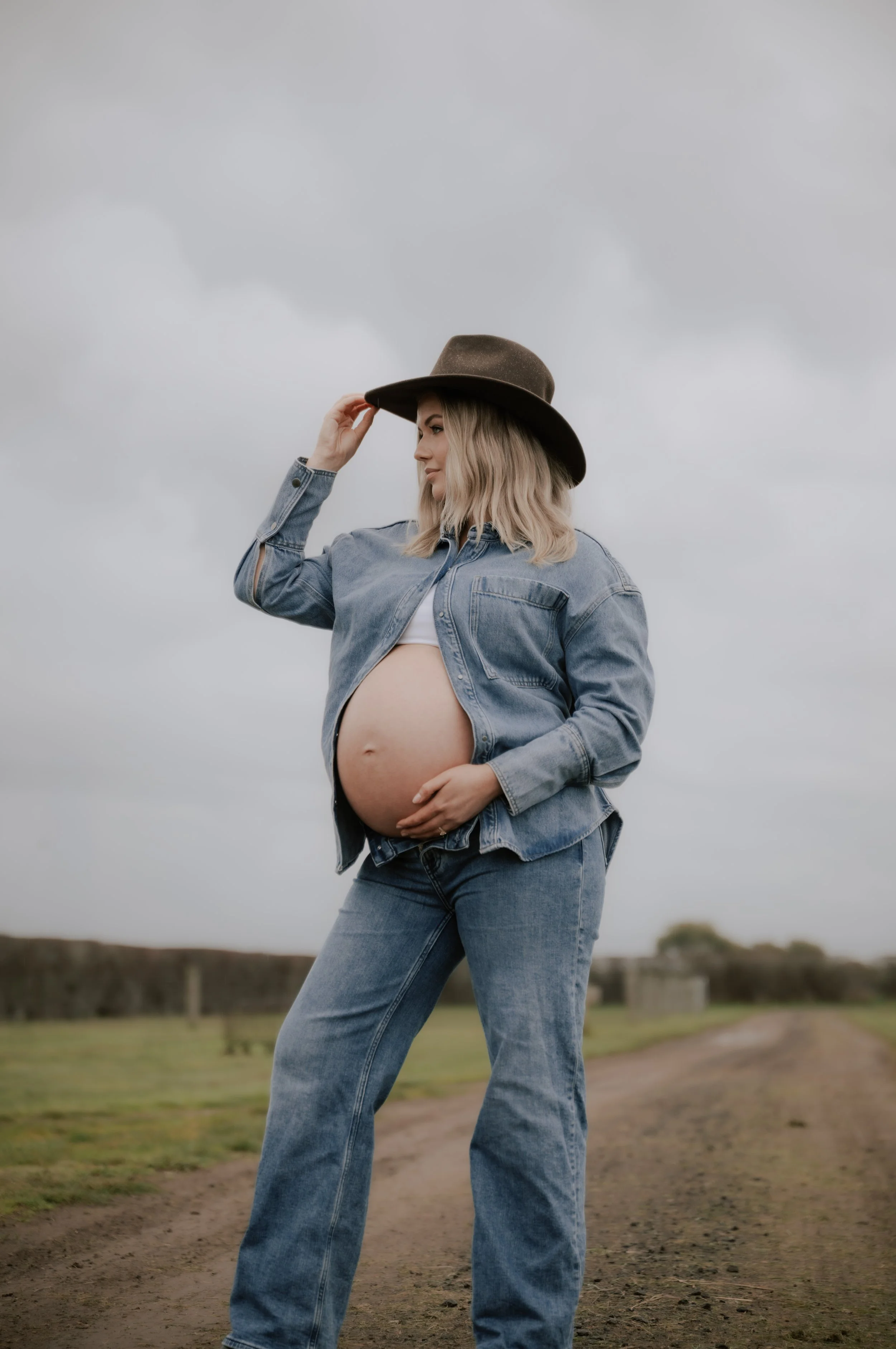 A pregnant woman in a denim jacket and jeans standing outdoors on a cloudy day, wearing a wide-brimmed hat, holding the brim with her right hand and resting her left hand on her belly.