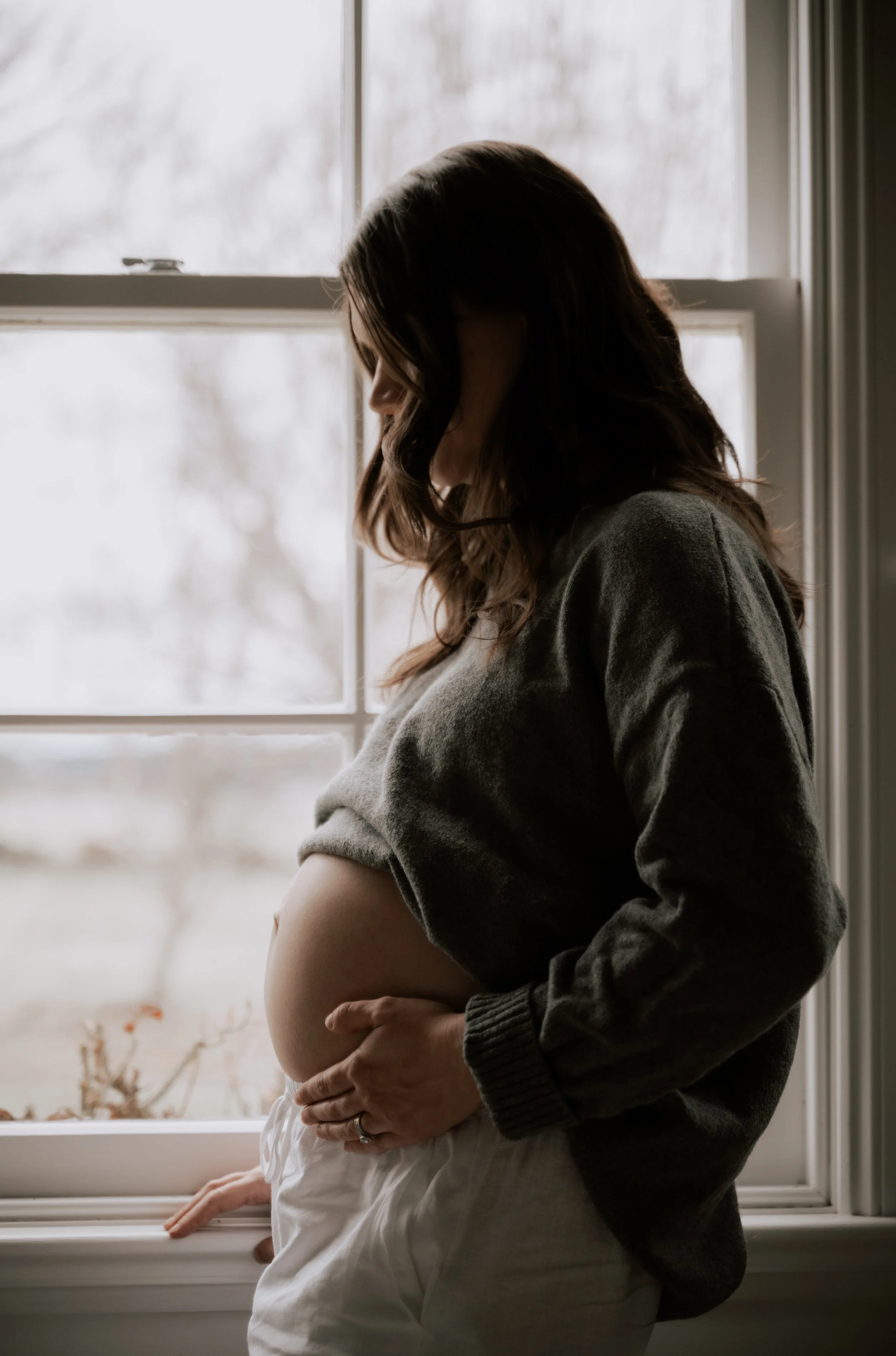 A pregnant woman in a gray sweater and a pregnant woman in white sitting by a window with bare trees outside visible in the background.
