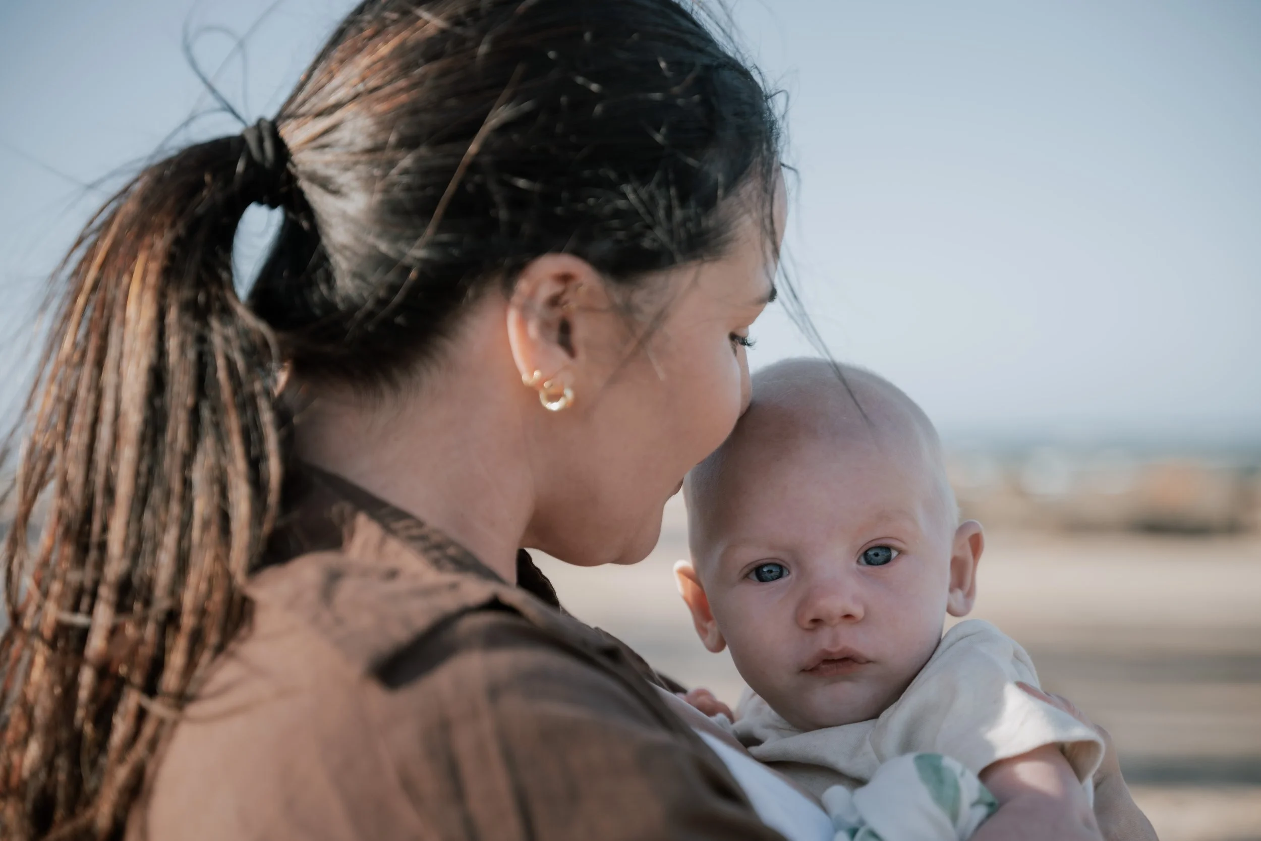 A woman with braided hair and earrings is cuddling a young child with blue eyes on a beach, with a clear sky in the background.