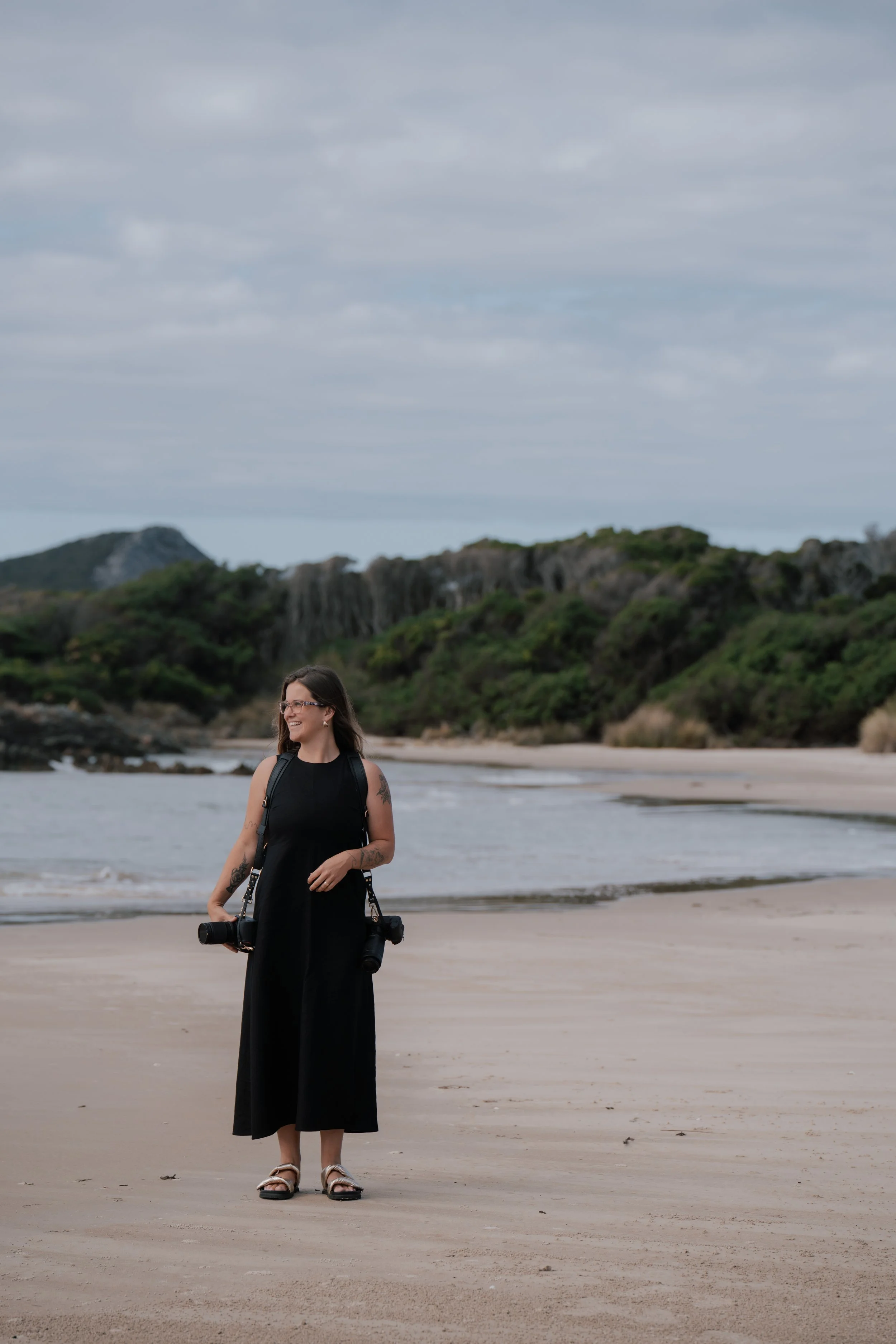 A wedding photographer, Mieke Lauren, wearing a black dress, sandals, and glasses standing on a sandy beach, holding two cameras with a scenic landscape of greenery, rocky hills, and cloudy sky in the background.