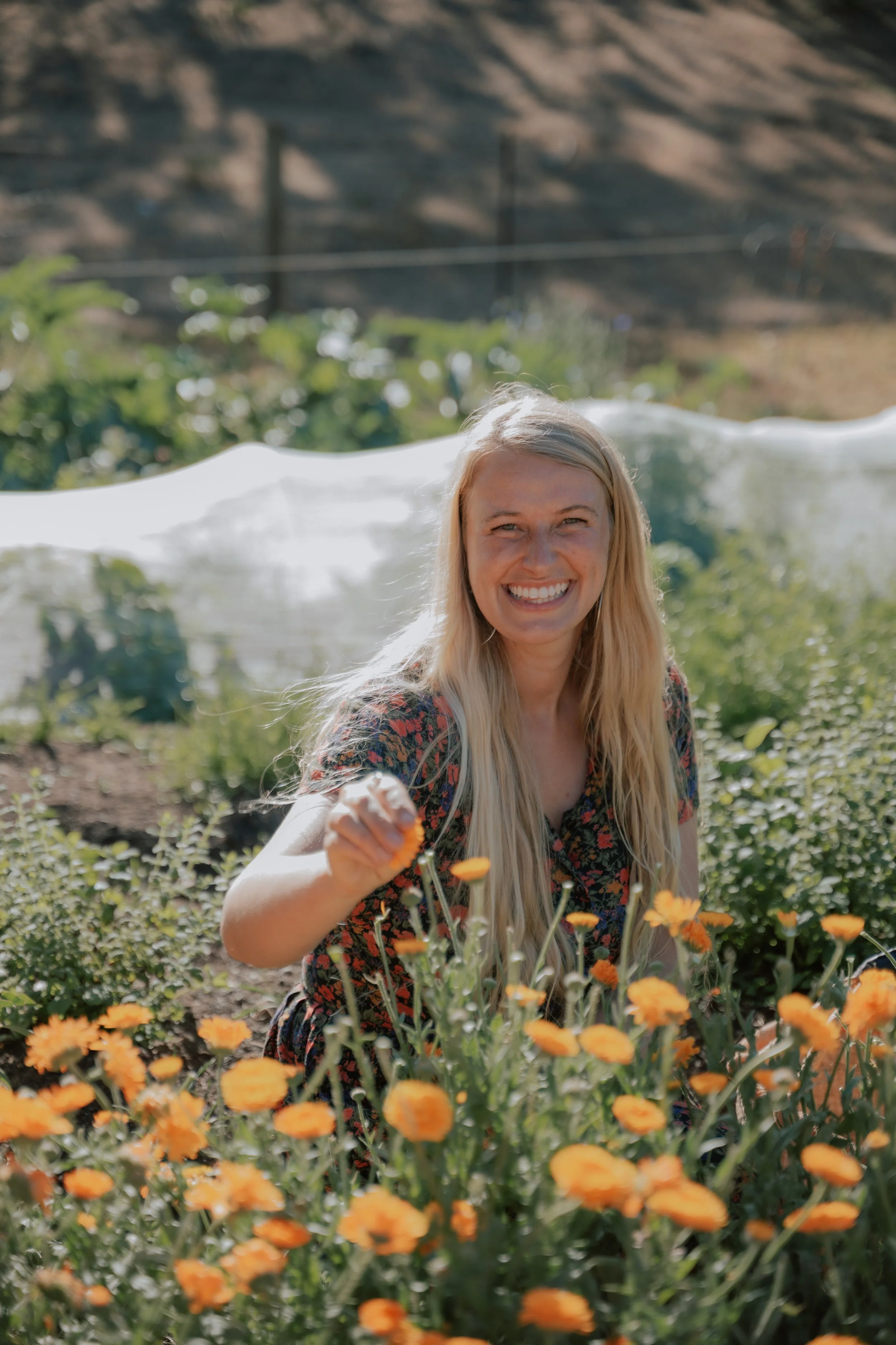 A smiling woman with long blonde hair in a garden surrounded by orange flowers, holding a flower in her hand.