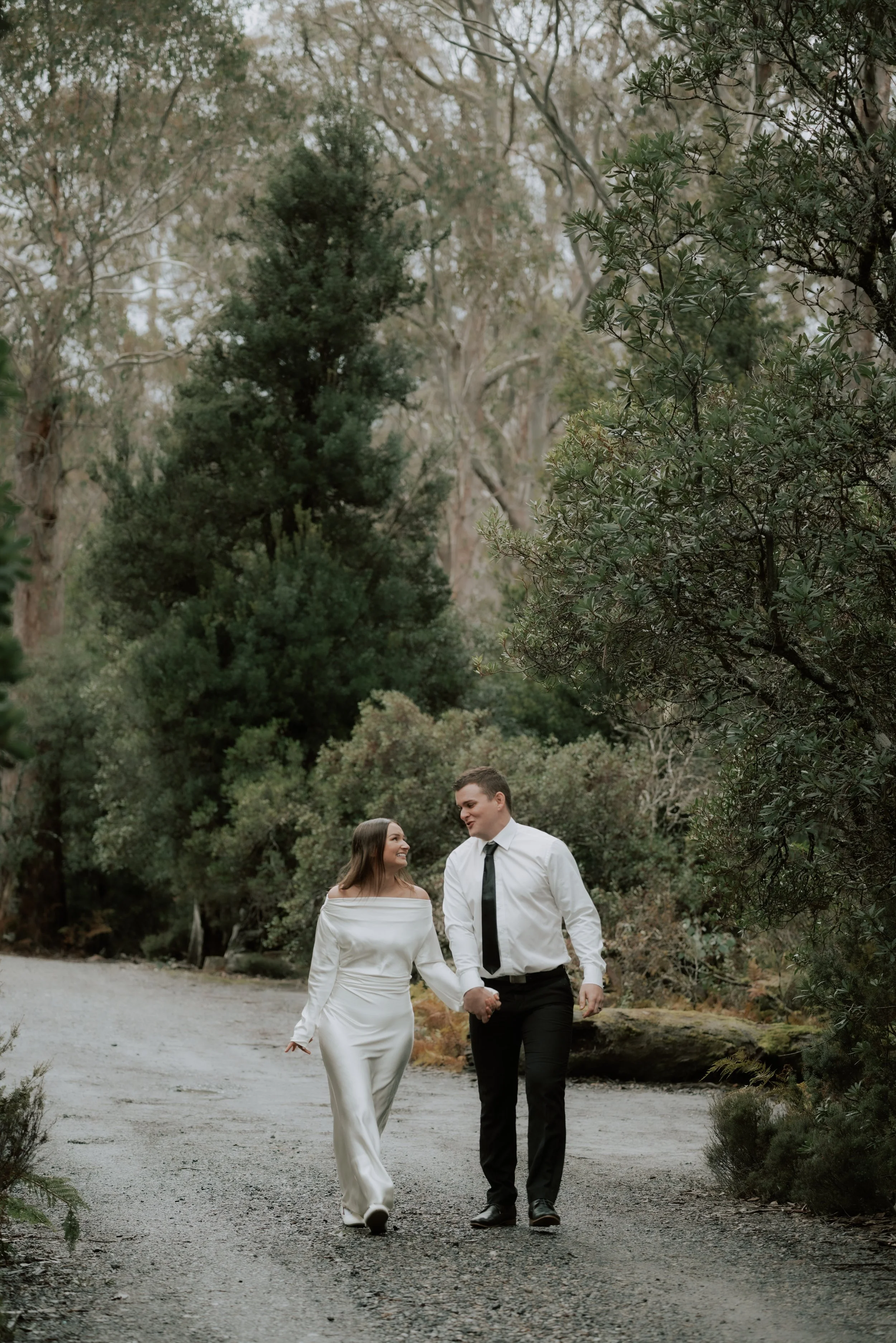 A couple walking hand in hand on a gravel path through a forest, holding hands and smiling at each other.