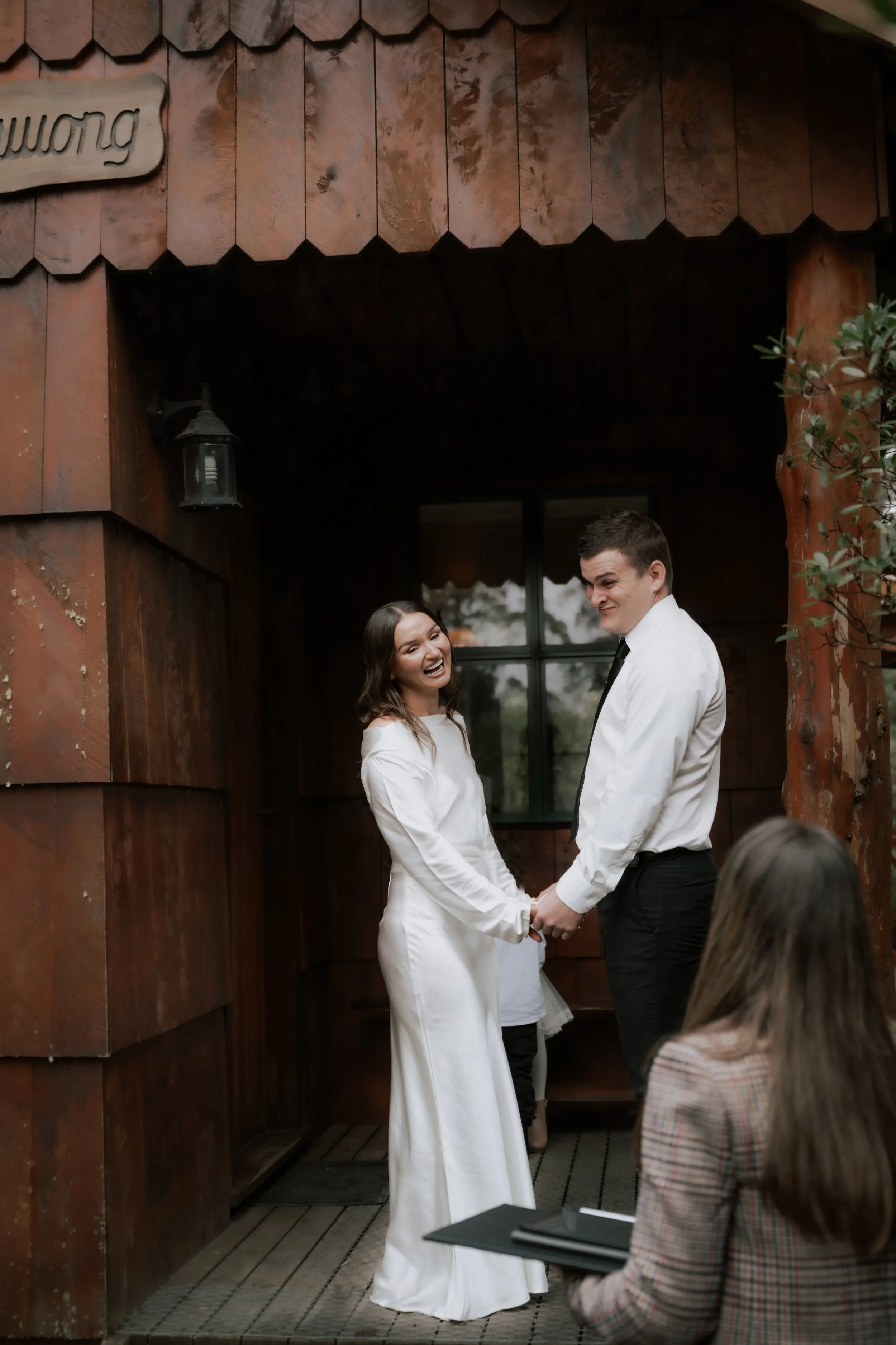A bride and groom holding hands during their wedding ceremony, standing outside in front of a wooden building with a window behind them. The bride is wearing a white wedding dress, and the groom is dressed in a white shirt and black tie, smiling at e