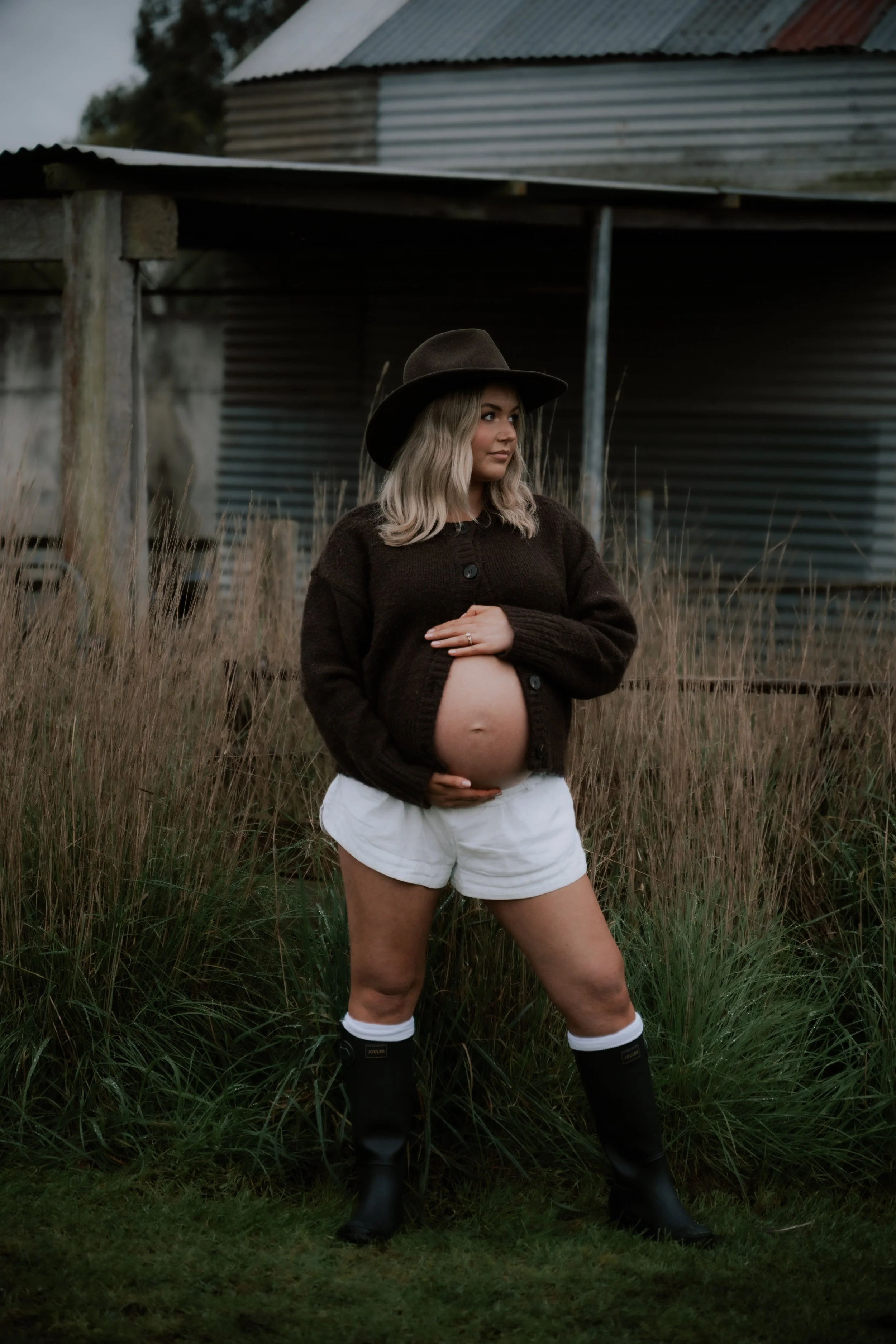 A pregnant woman with blonde hair wearing a black hat, a black cardigan, white shorts, and rain boots, standing outdoors in tall grass with an old wooden and metal building in the background.